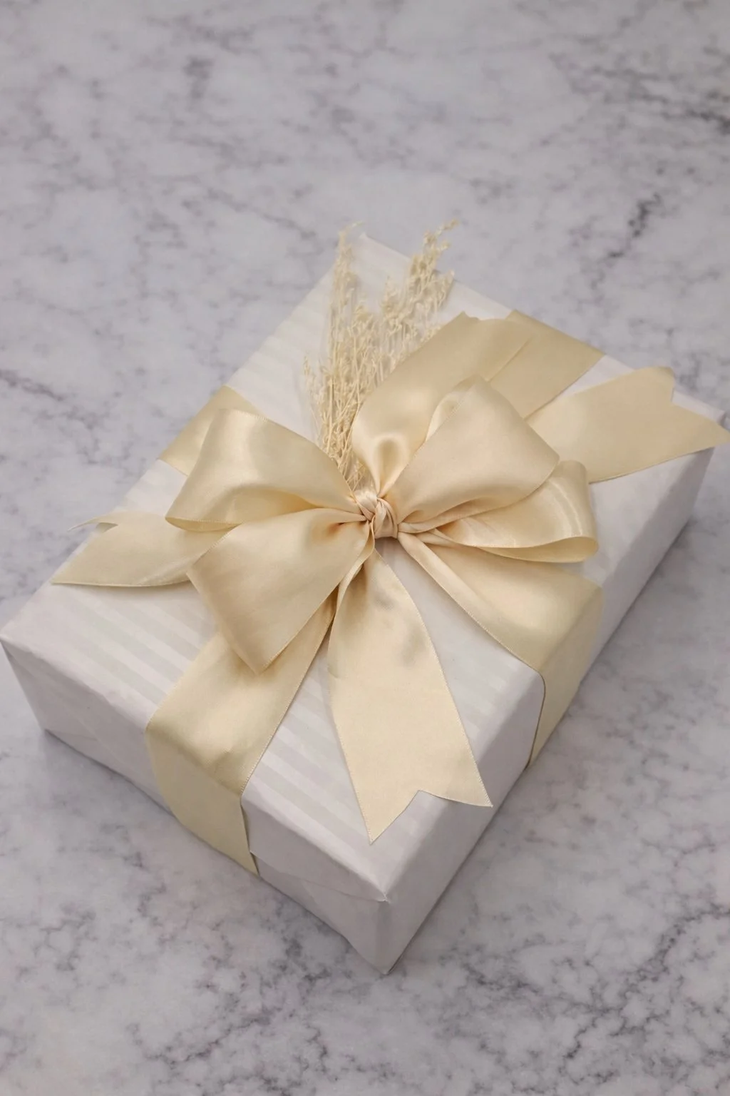 A gift wrapped in white paper with a gold satin ribbon tied in a bow, placed on a gray marble surface.