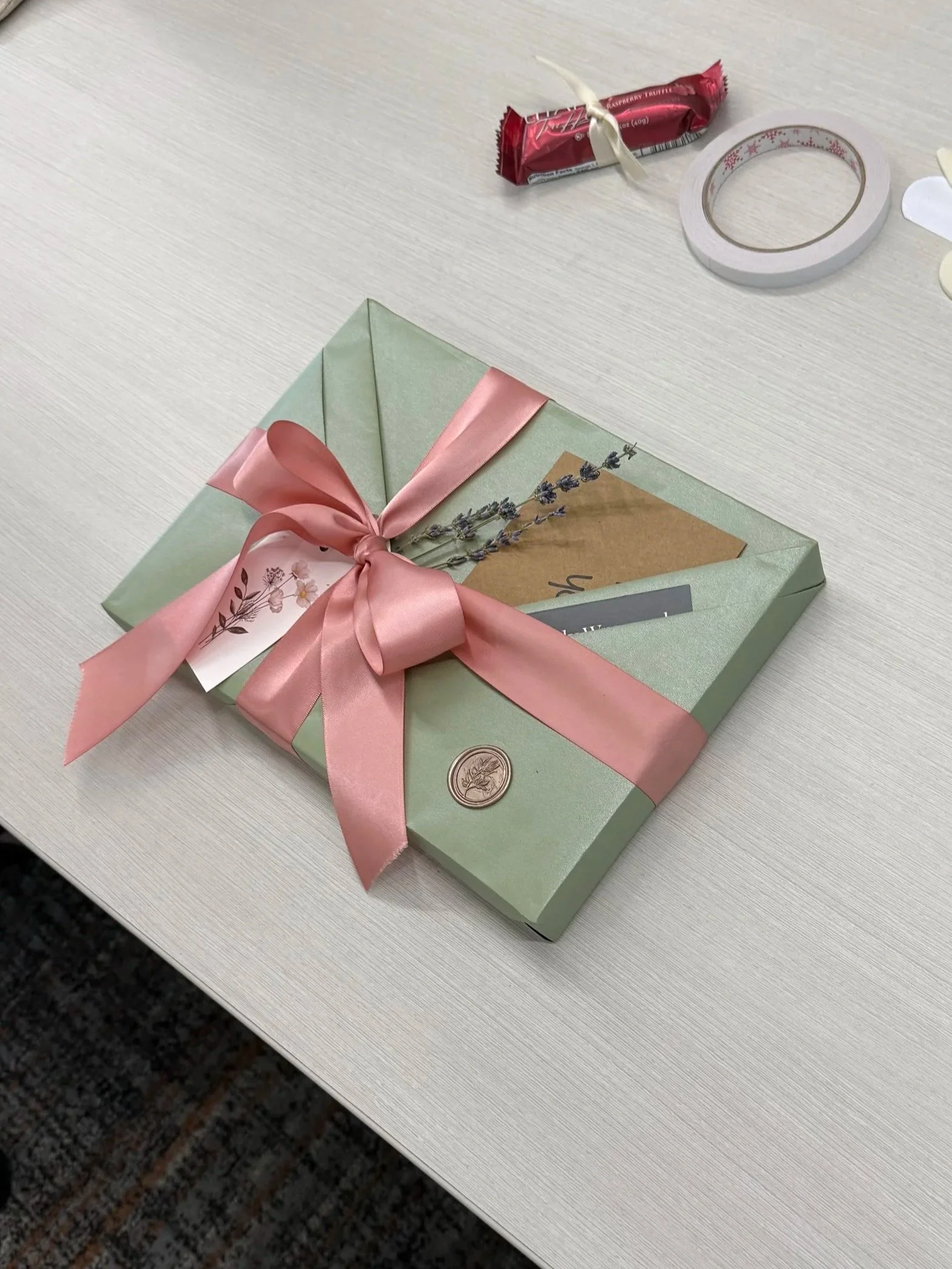 A gift box wrapped in light green paper with a pink satin ribbon bow on top, decorated with sprigs of lavender, on a white table.