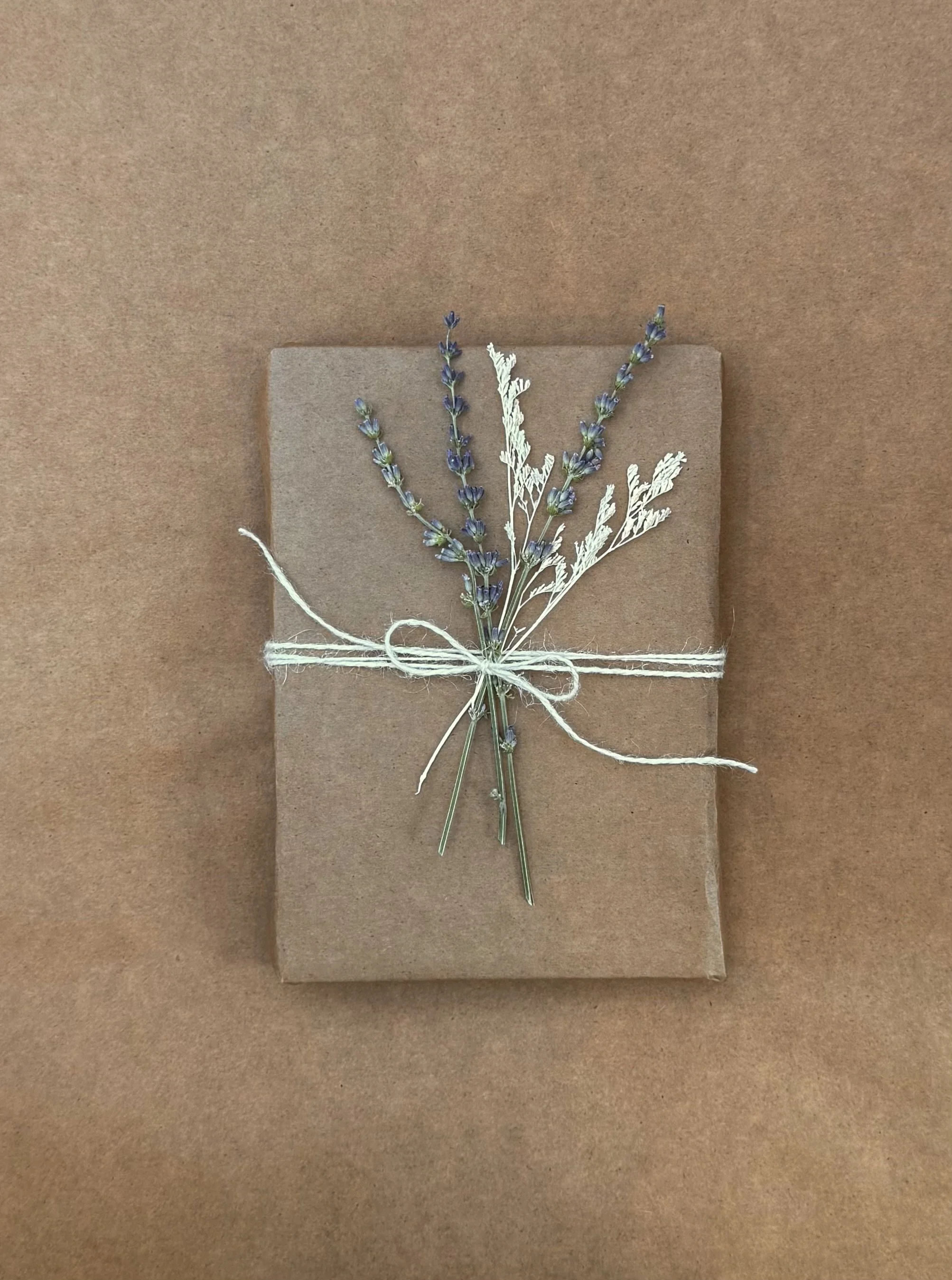 A gift wrapped in plain brown paper, decorated with sprigs of lavender and dried white foliage, tied with white string.