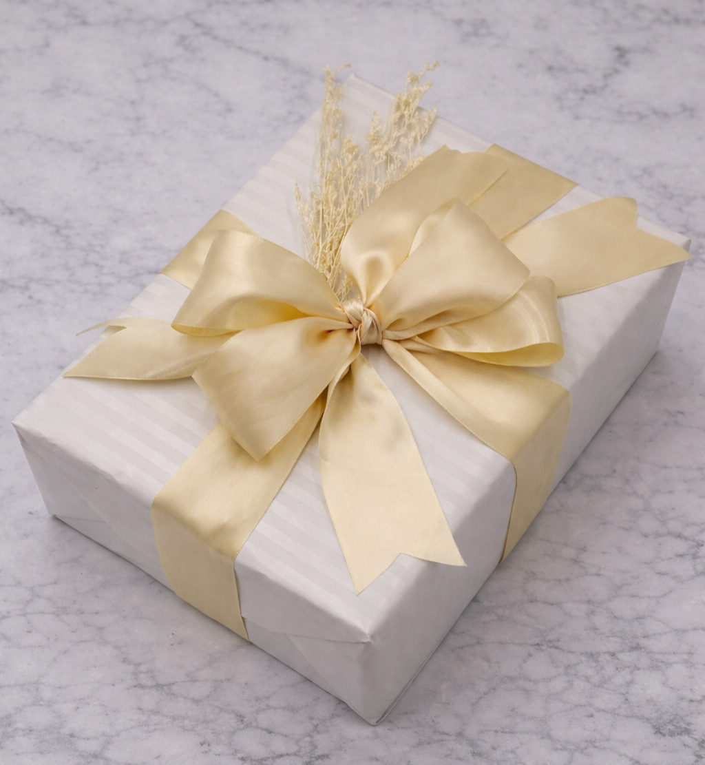 Decorative gift box wrapped in white paper with a gold satin ribbon tied in a bow and a small dried flower arrangement on top, placed on a marble surface.
