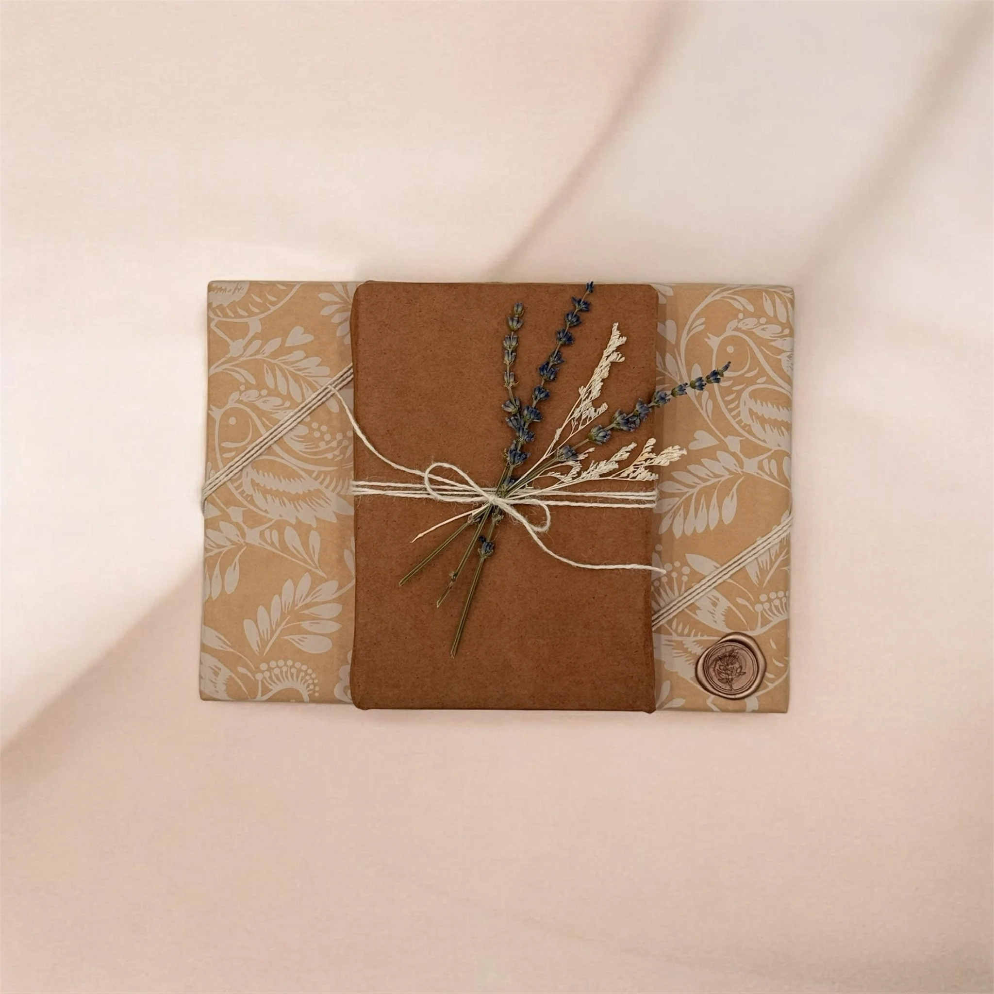 A wrapped gift with a brown paper package tied with string, decorated with dried lavender and wheat sprigs, placed on top of another gift wrapped in beige paper with white floral and leaf patterns, sealed with a wax stamp.