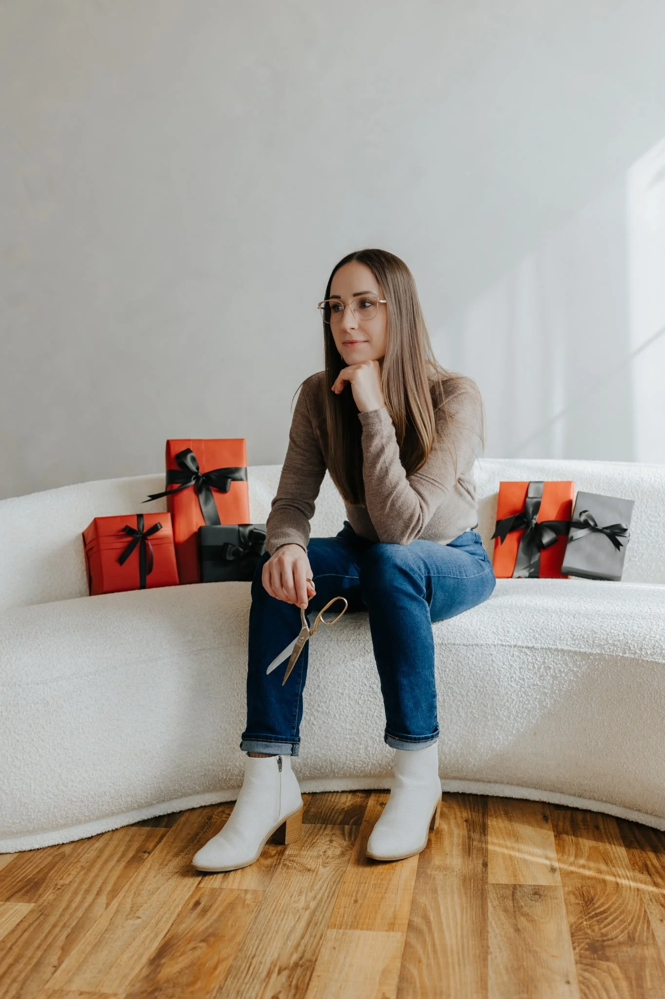 A woman sitting on a white sofa with a pair of scissors in her hand, surrounded by wrapped gift boxes with ribbons, in a bright room with wooden flooring.