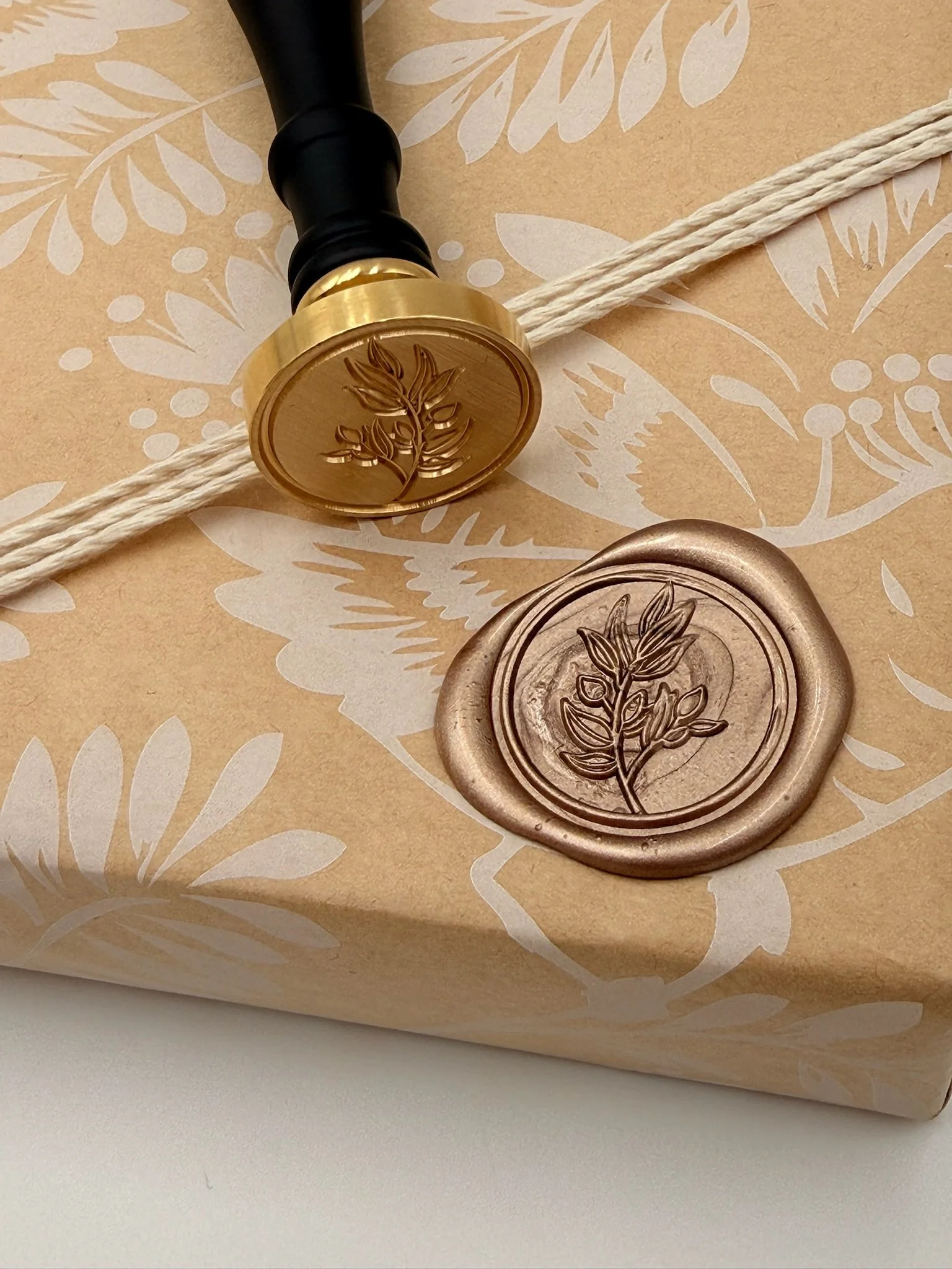 Gold and rose-colored wax seals with embossed leaf design, one sealing a wrapped gift with decorative paper, a black handle stamp, and a cream-colored string.