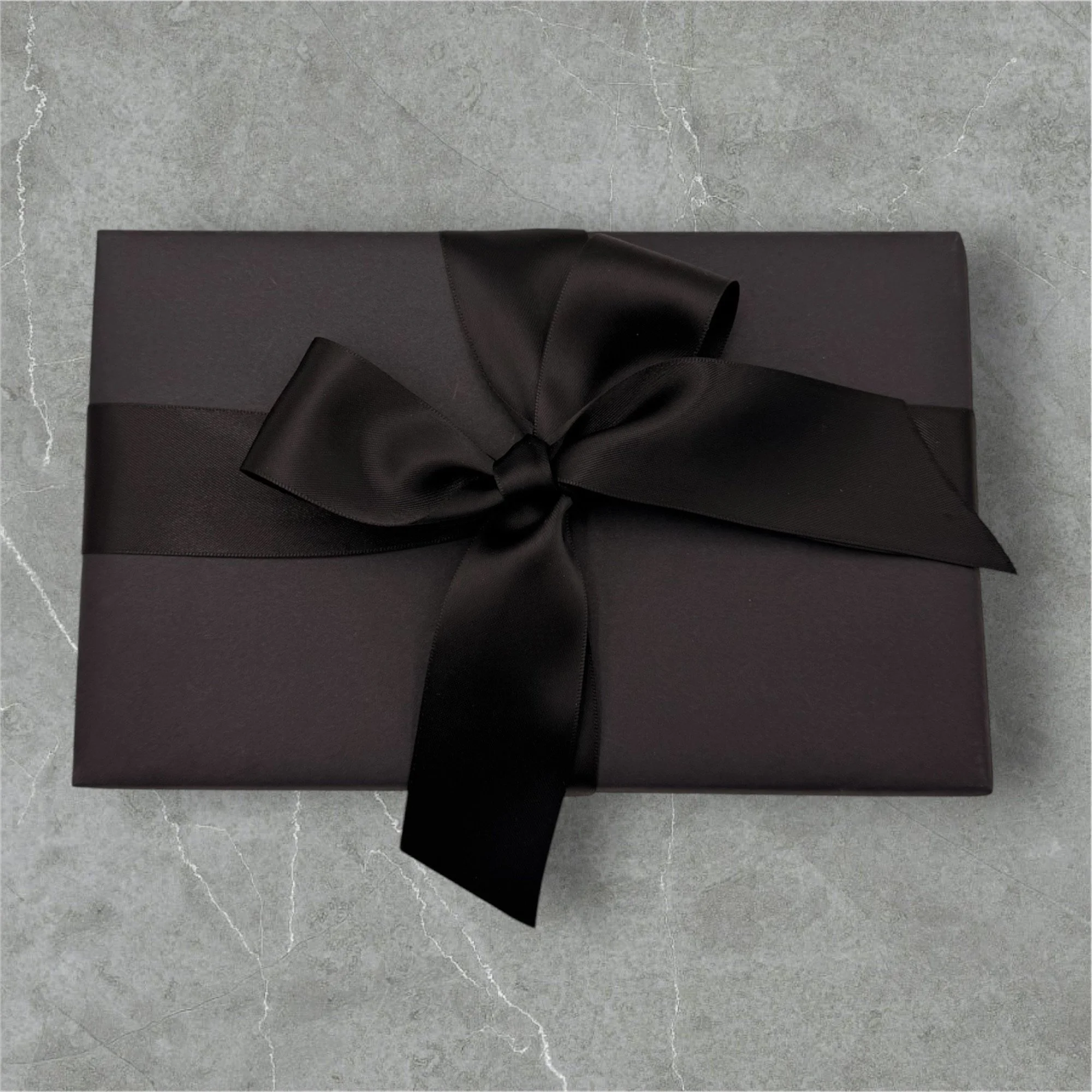 A rectangular black gift box wrapped with a black satin ribbon tied in a bow on top, placed on a gray marble surface.