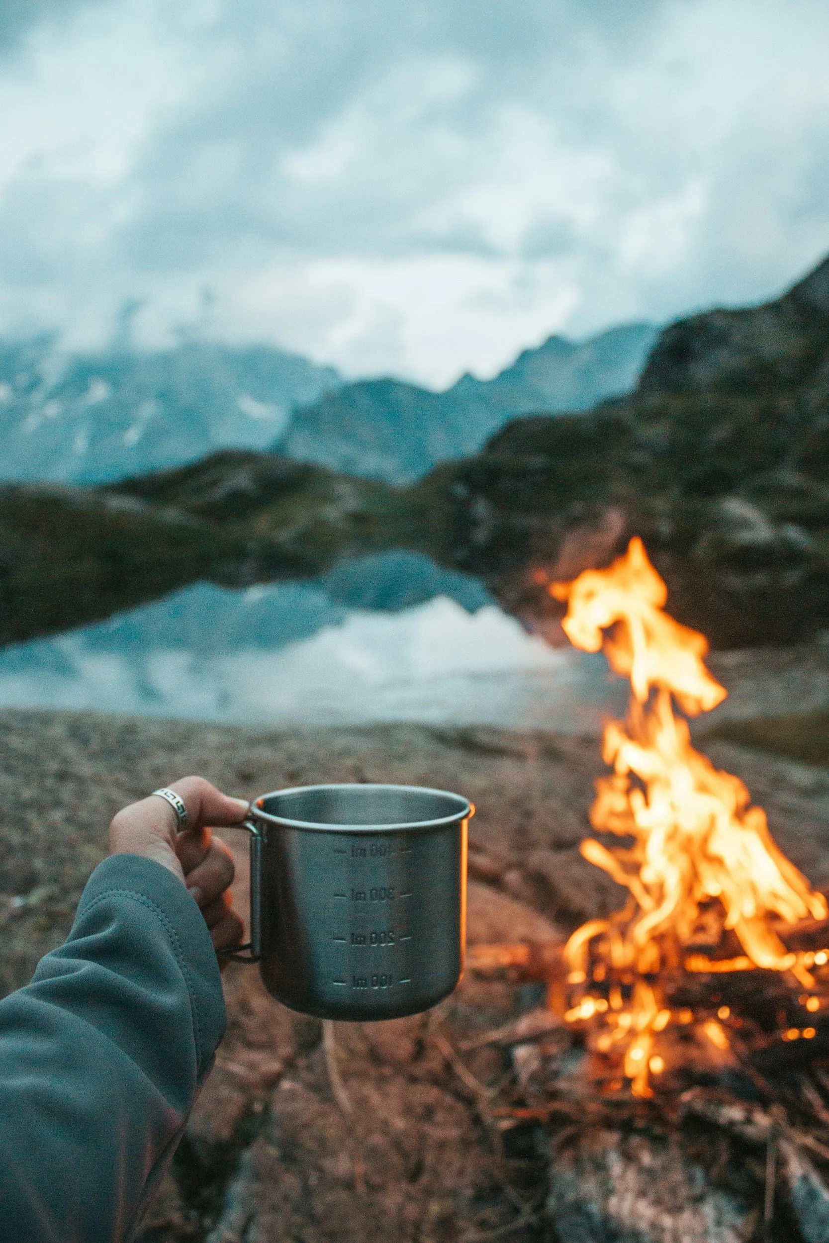 A person holding a metal measuring cup near a campfire with mountains and a cloudy sky in the background.