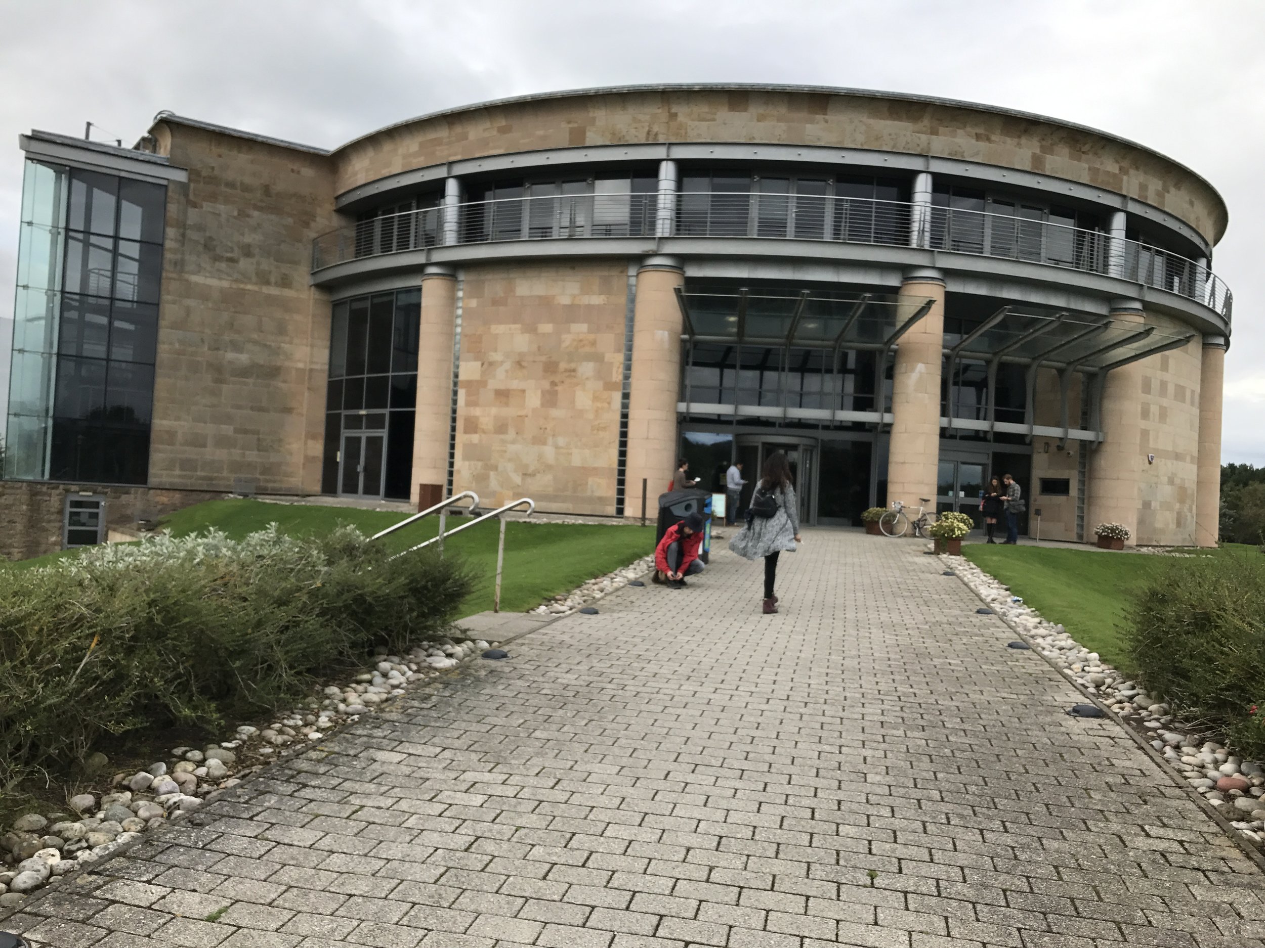 A modern building with curved architecture, large glass windows, and a stone facade, with people walking on the paved pathway leading to the entrance.