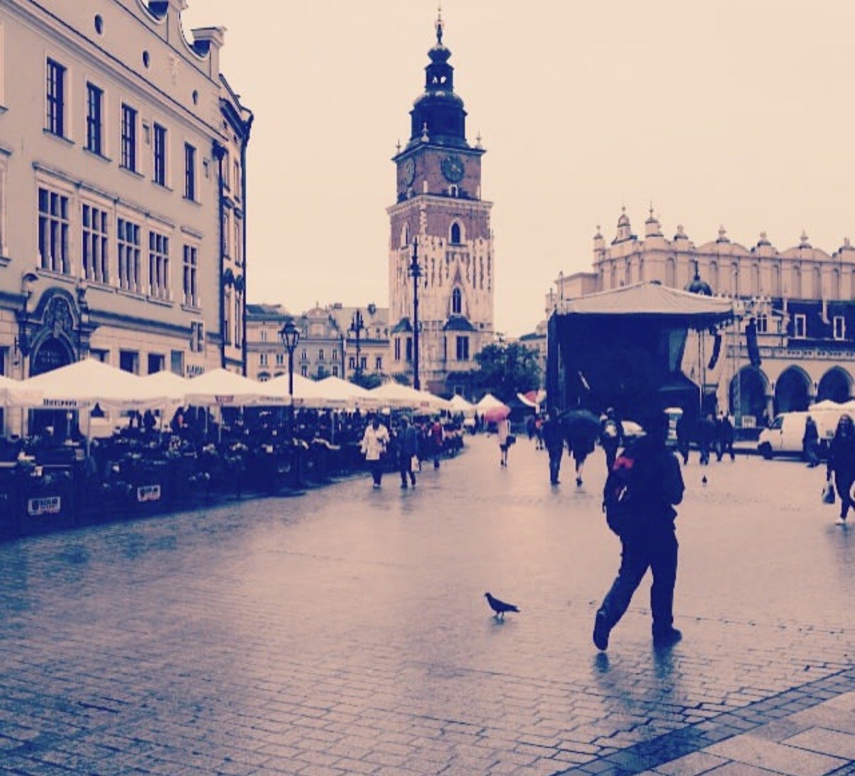 A lively European town square with historic architecture, outdoor cafes, a clock tower, and people walking on cobblestone streets.
