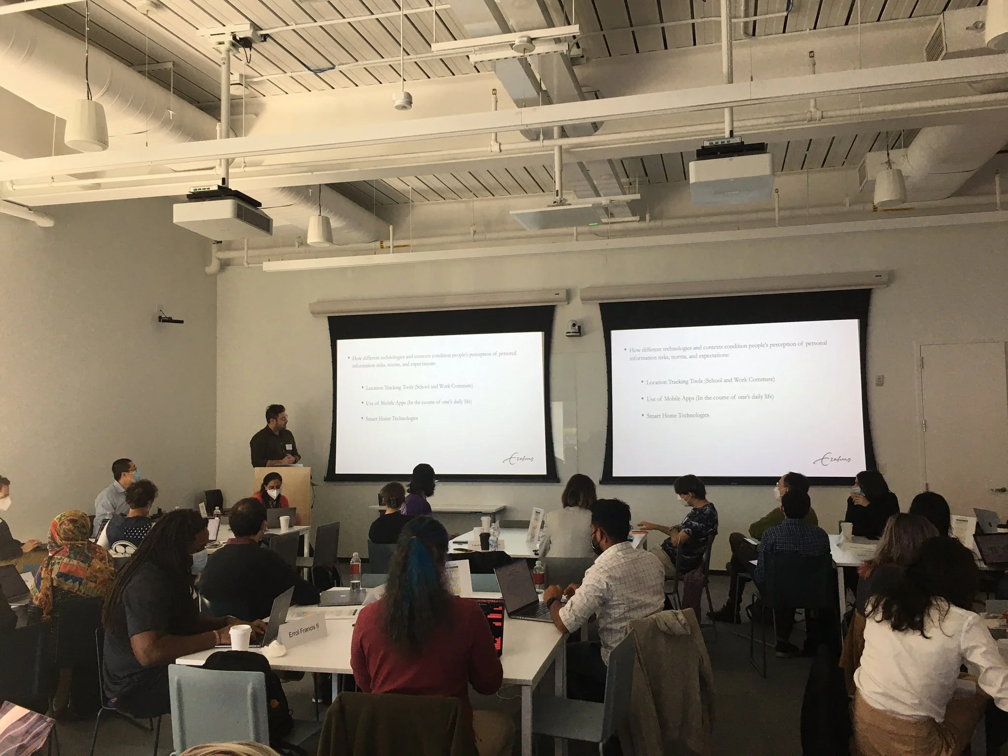 People attending a conference or seminar in a modern classroom, some wearing face masks, with two large presentation screens at the front displaying a slide about technology and personal safety.