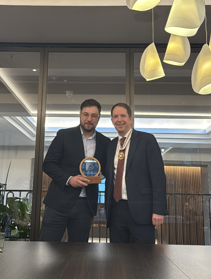 Two men in suits standing inside a modern office or conference room, one holding a trophy or award, smiling for the camera with a large window behind them and contemporary light fixtures above.