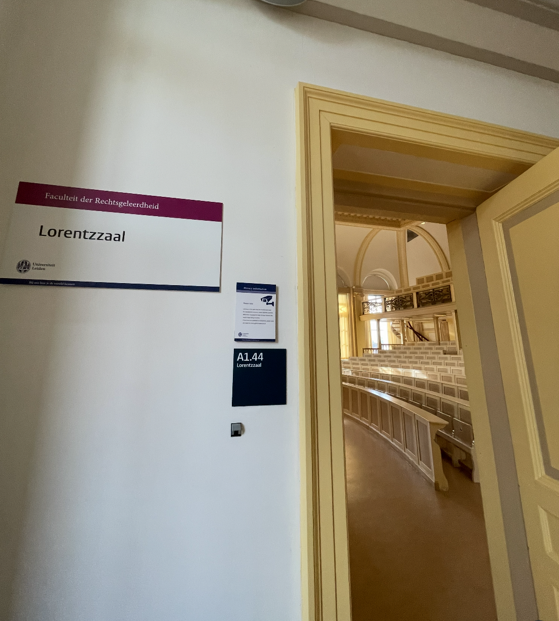 Sign on a wall indicating Lorentzaal in the Faculty of Law at Leiden University, with an open door leading into an auditorium or lecture hall with rows of seating and decorative interior details.