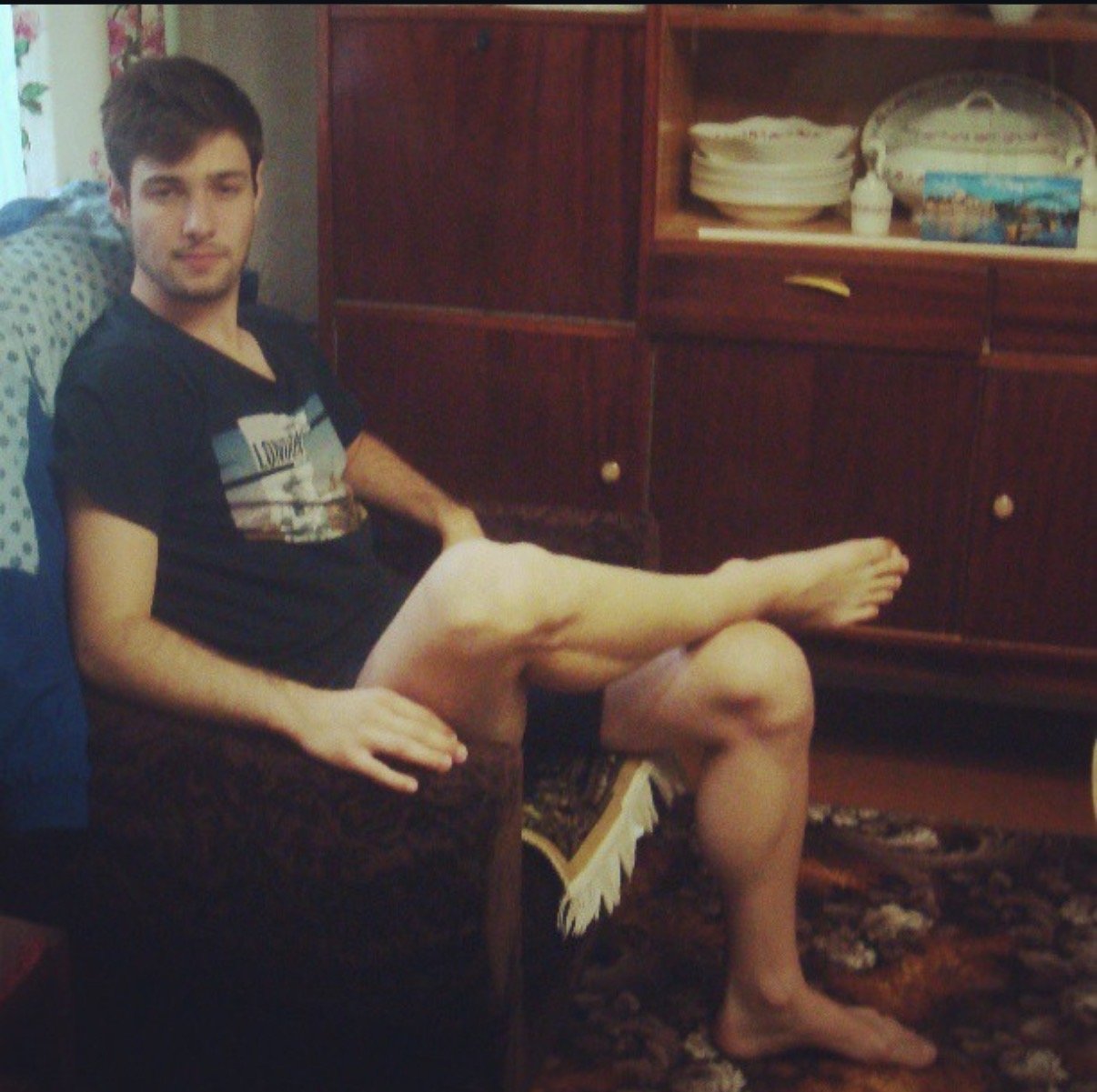 A young man sitting on a chair in a living room, with his right leg crossed over his left knee and his left arm resting on the armrest. He is wearing a black T-shirt and has short brown hair. Behind him, there is a wooden cabinet with dishes and decorations on top, and a patterned carpet on the floor.