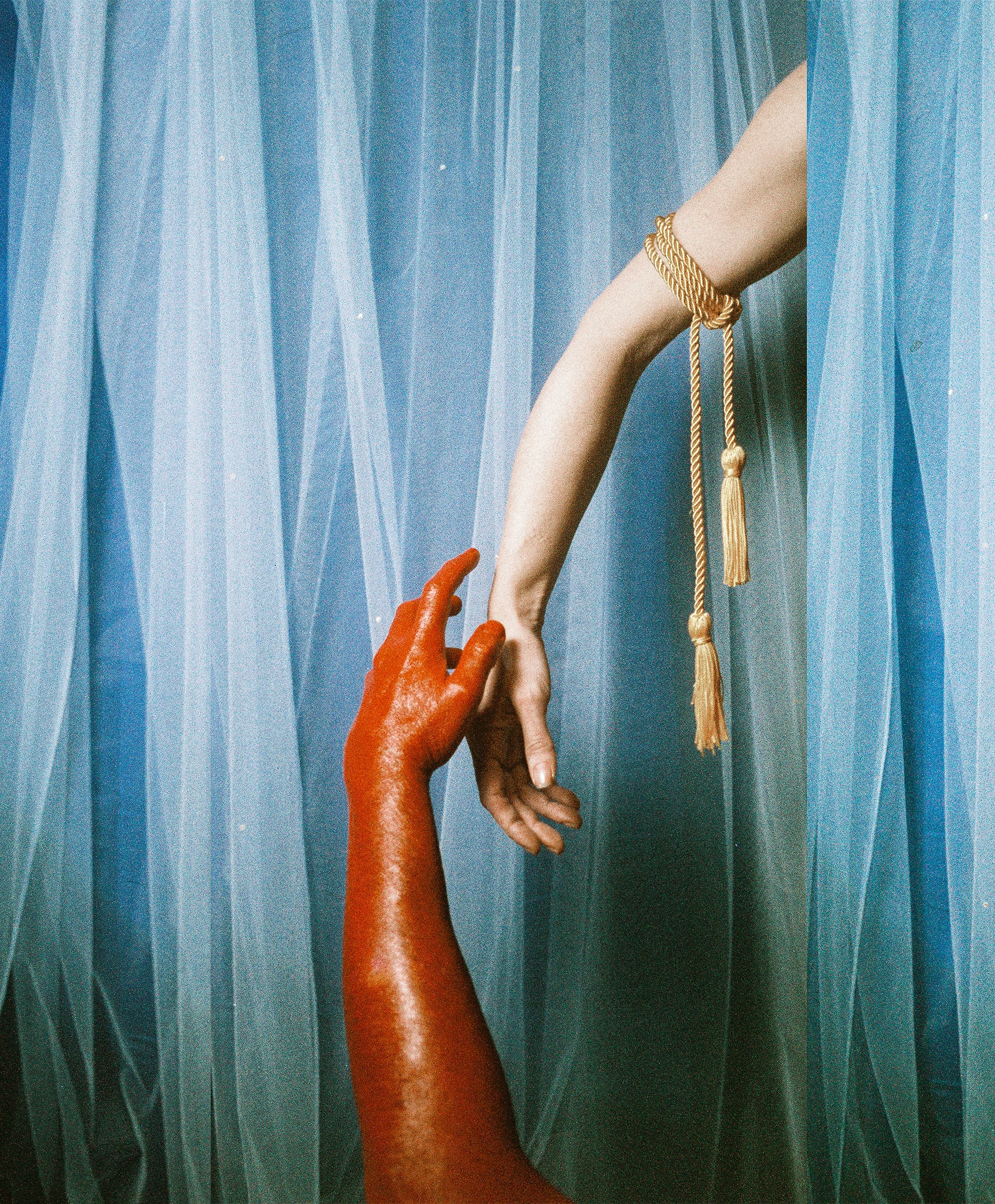 Two hands reaching towards each other against a textured blue background. One hand is painted red and the other is natural skin tone with a gold bracelet.