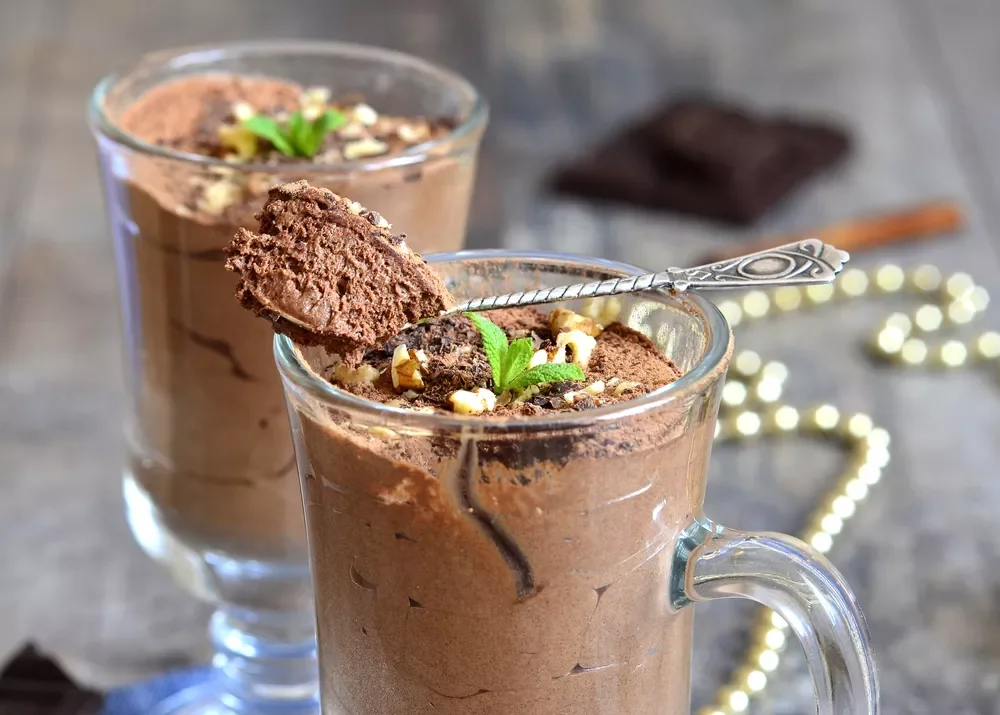 Deux verres de mousse au chocolat garnis de chocolat râpé, de morceaux de noix, et de feuilles de menthe, avec une cuillère en argent insérée dans un des verres.