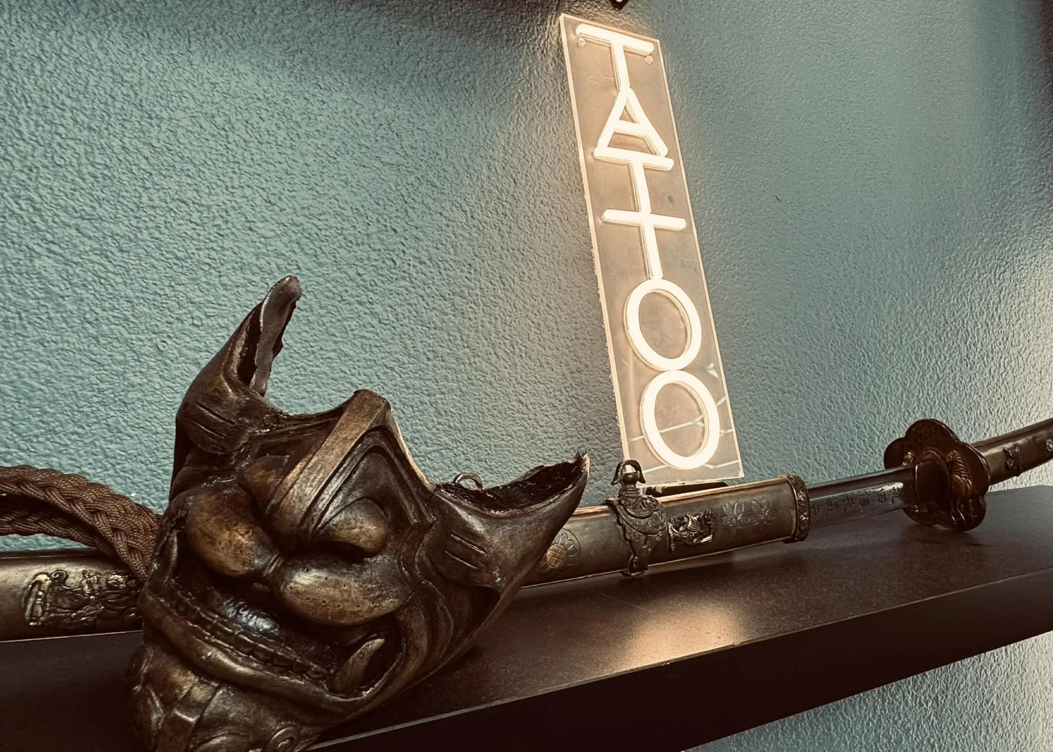Decorative samurai sword with a hand sculpted samurai mask. art decor for a tattoo parlor/ studio.  In the background a tattoo neon sign.