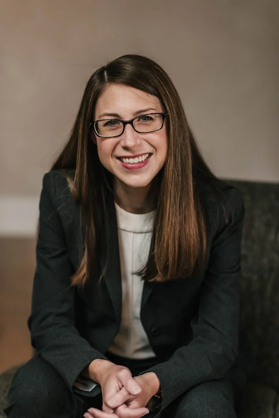A young woman with long brown hair, wearing glasses and a dark blazer, smiling while sitting indoors.