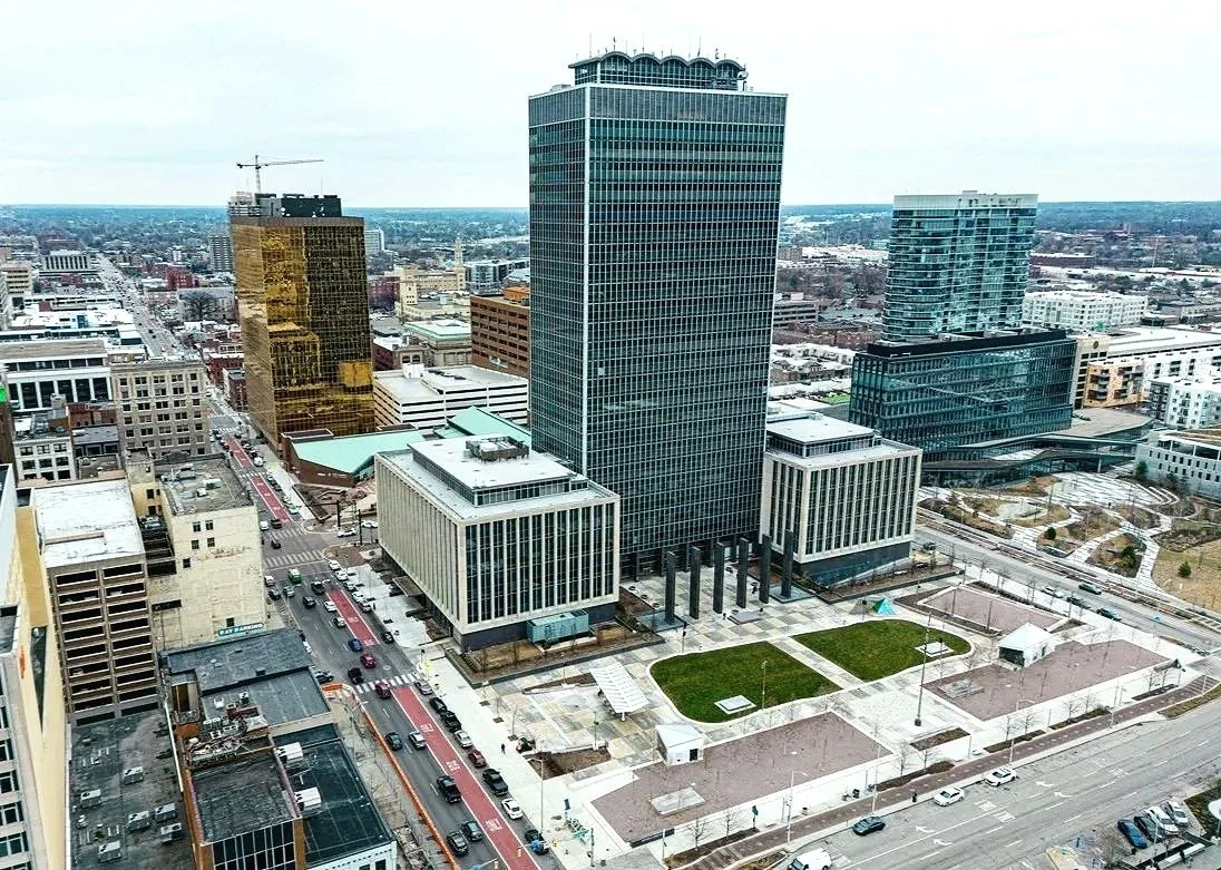 Aerial view of downtown city buildings, including a tall office tower with a rounded top, modern glass structures, and streets with cars and pedestrians.