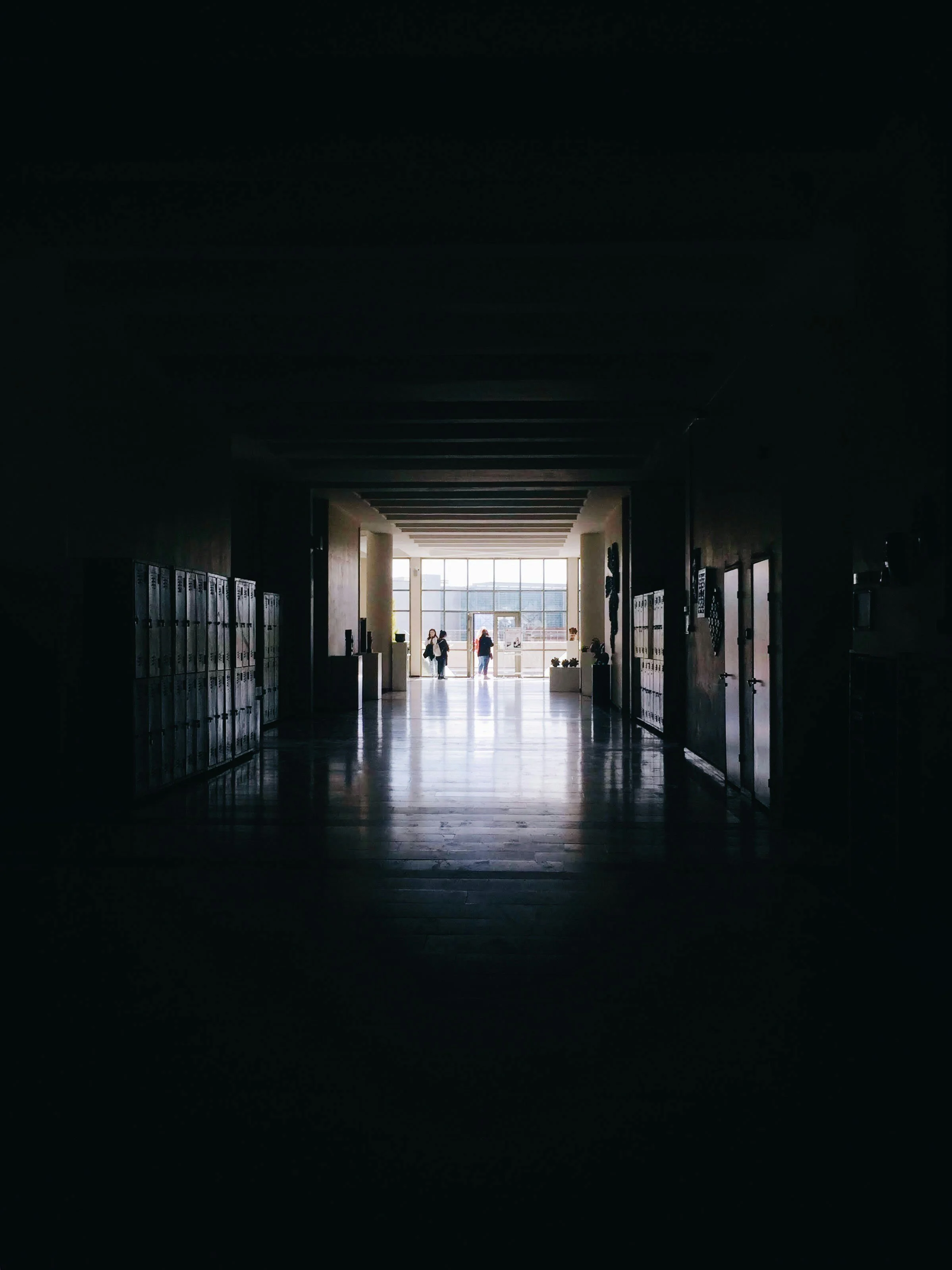 School hallway with lockers on the sides, leading to a brightly lit entrance with large windows. Several students are visible near the entrance.