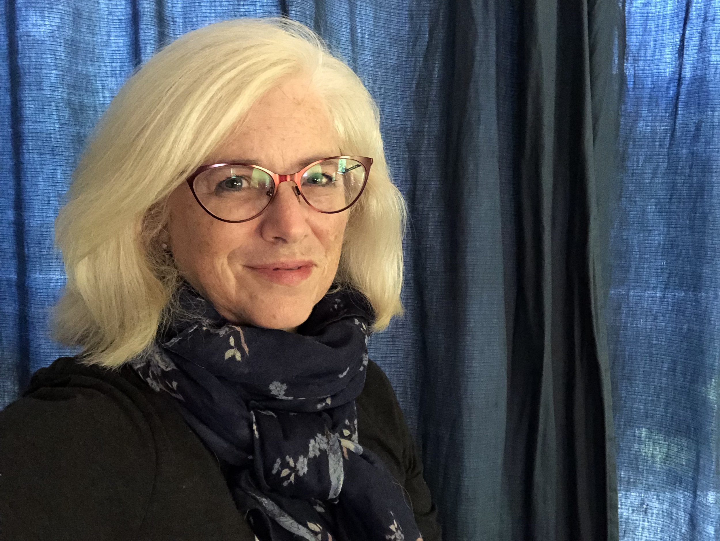 A woman with blonde hair wearing glasses and a dark scarf smiles in front of dark blue curtains.