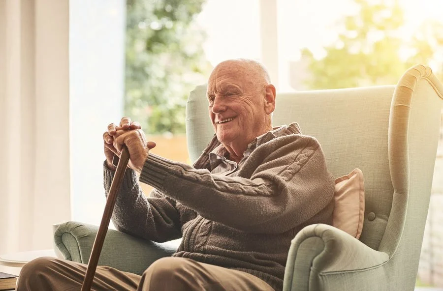 An elderly man sitting in a comfortable armchair, smiling and holding a walking cane, with a sunny window behind him.