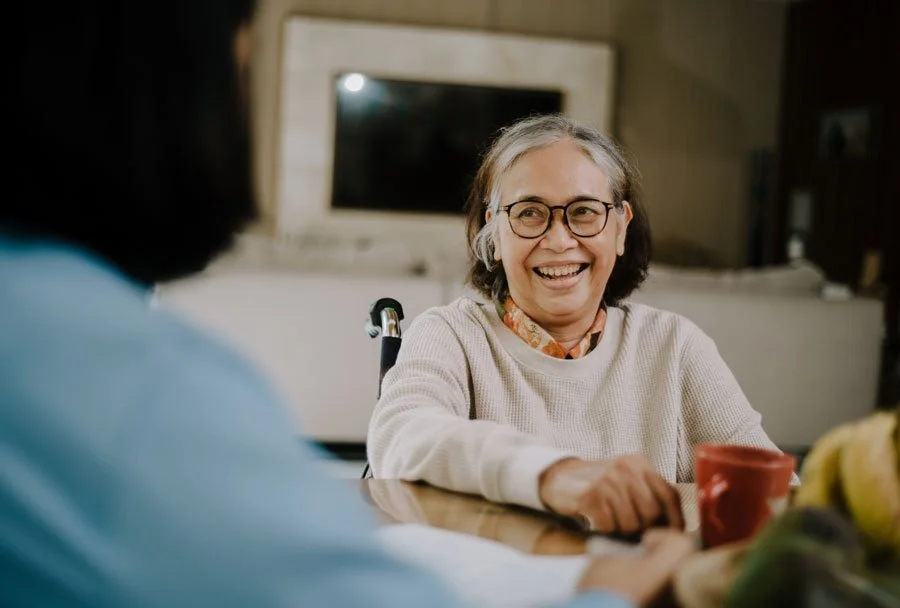 An elderly woman with glasses smiling and talking to another person at a table, with a red mug and bananas on the table.