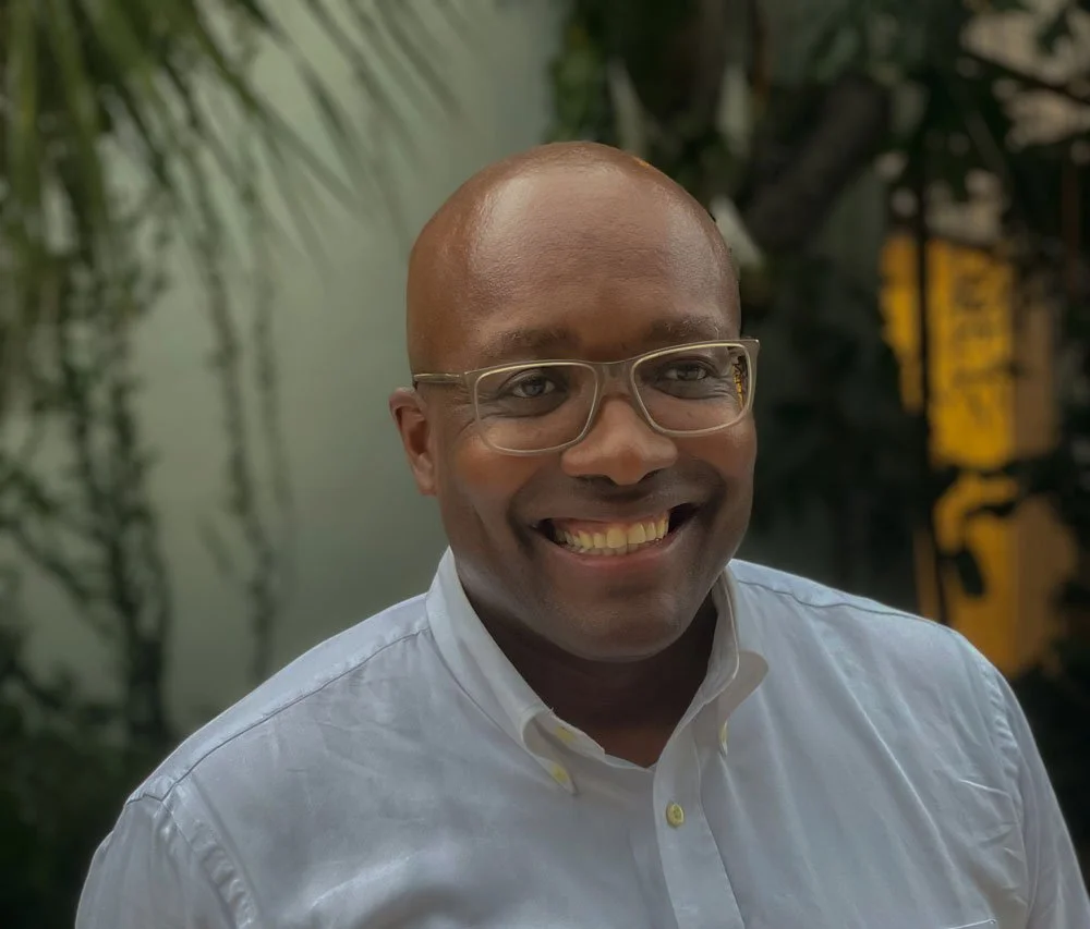 A man with glasses and a white button-up shirt smiling at the camera, standing outdoors with green plants in the background.