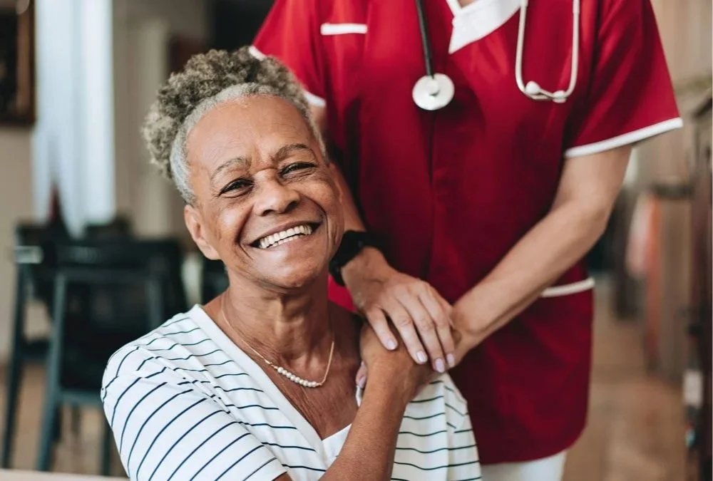 A joyful elderly woman with gray curly hair smiling at a healthcare worker in red scrubs. The healthcare worker is gently holding her hand.
