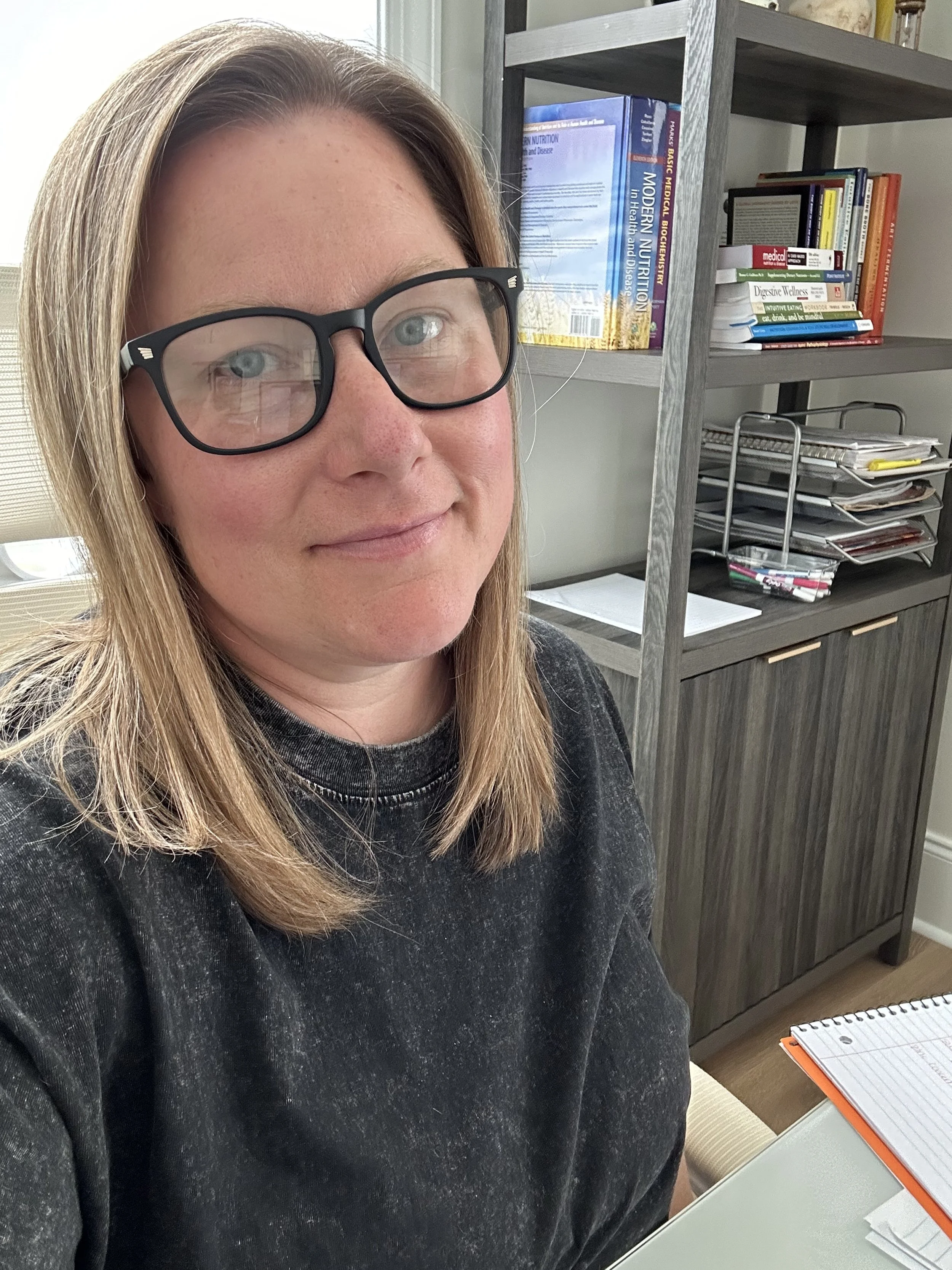A woman with shoulder-length blonde hair, glasses, and a black shirt sitting at a desk with a spiral notebook, in an office with shelves filled with books and papers in the background.