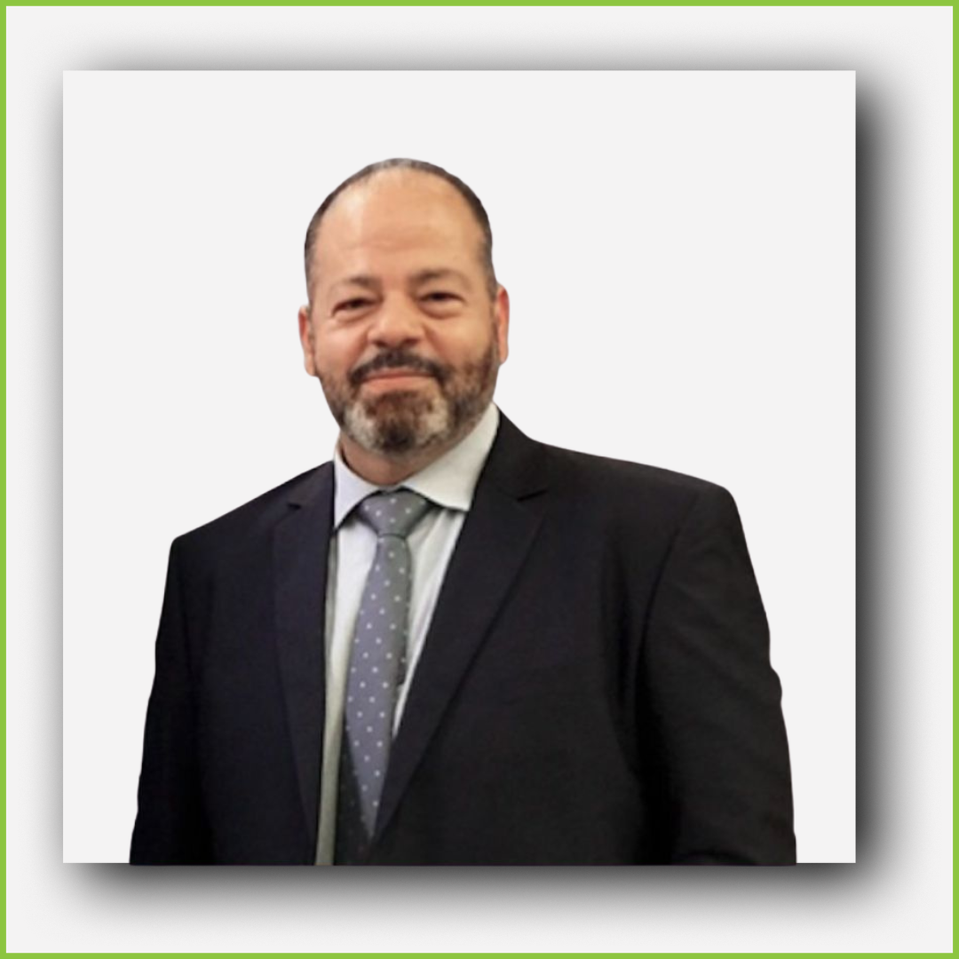 A man with a beard and receding hairline wearing a black suit, white shirt, and dotted gray tie, standing against a plain light background.