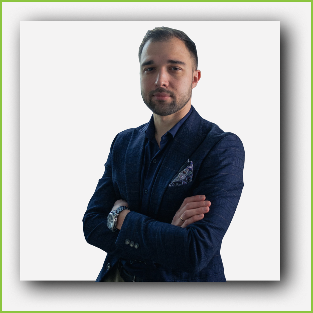 A young man with dark hair and beard, wearing a navy blazer, dark shirt, and pocket square, standing with arms crossed against a white background.