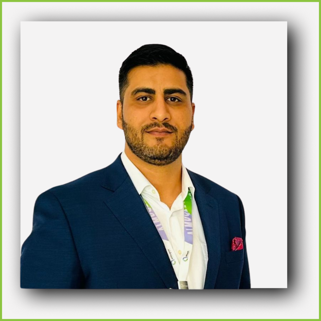 A man with dark hair and a beard, wearing a dark blue suit jacket, white shirt, and a lanyard, standing against a plain background.