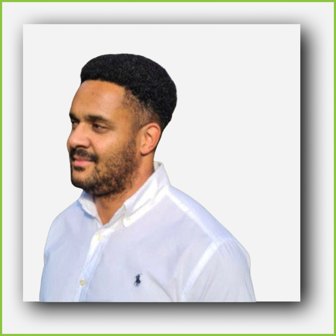 A man with short curly black hair, a beard, and mustache wearing a white collared shirt with a small black logo on the chest, standing against a plain light background.