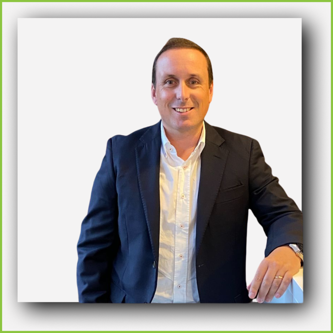 A man in a dark suit jacket and white shirt, smiling and posing with his hand resting on a surface against a plain white background.