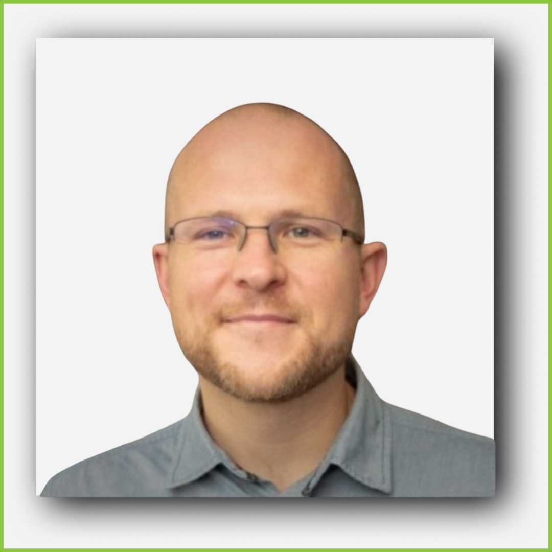 Headshot of a man with glasses, light beard, wearing a gray button-up shirt, smiling softly, against a plain light background.
