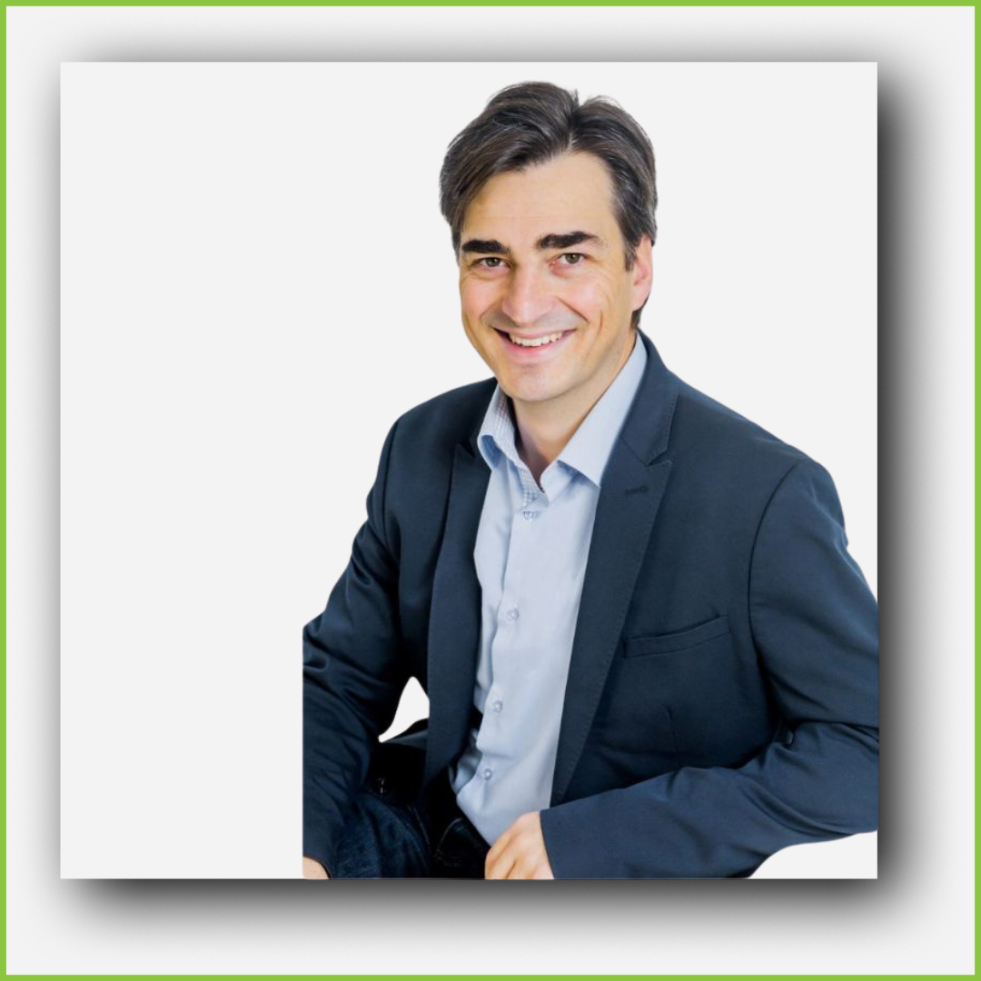 A man with dark hair in a blue suit and light blue shirt smiling, sitting with one arm resting on a chair.