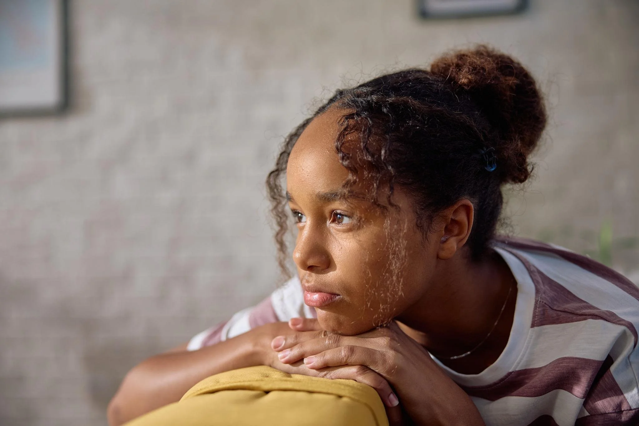 Young woman with curly hair in a bun, resting her chin on her arms on a yellow surface, looking thoughtfully to the side, with a blurred background.