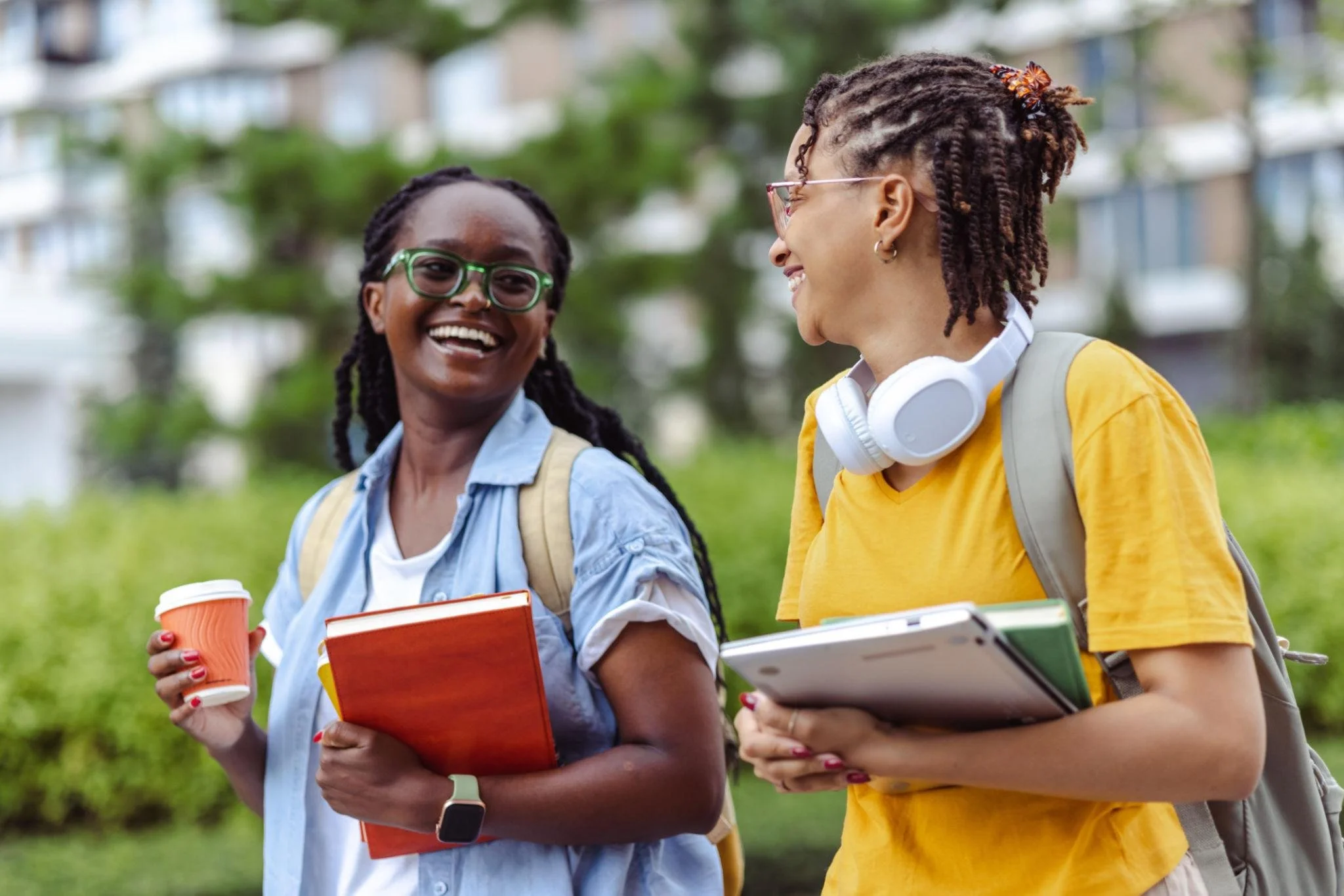 Two smiling women with glasses and backpacks walking outdoors, holding books and coffee cups, engaged in conversation.