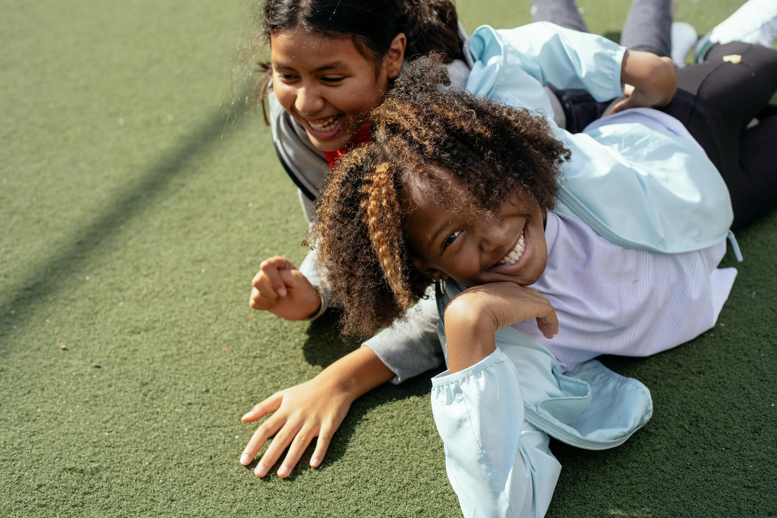 Two young girls are playing on the grass, laughing and lying intertwined with each other, enjoying each other's company outdoors.