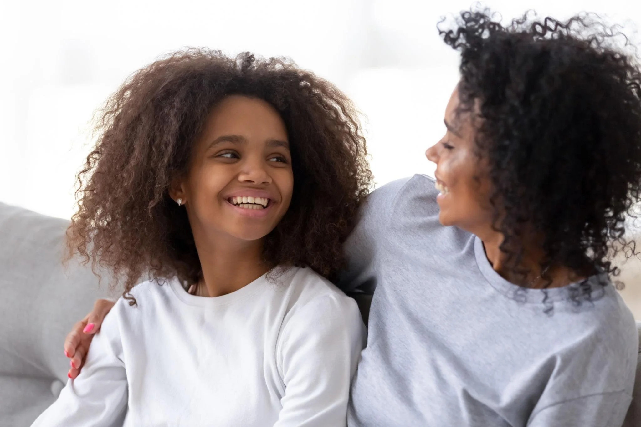 A woman and a girl with curly hair smiling and looking at each other, sitting close together on a light-colored couch in a bright room.