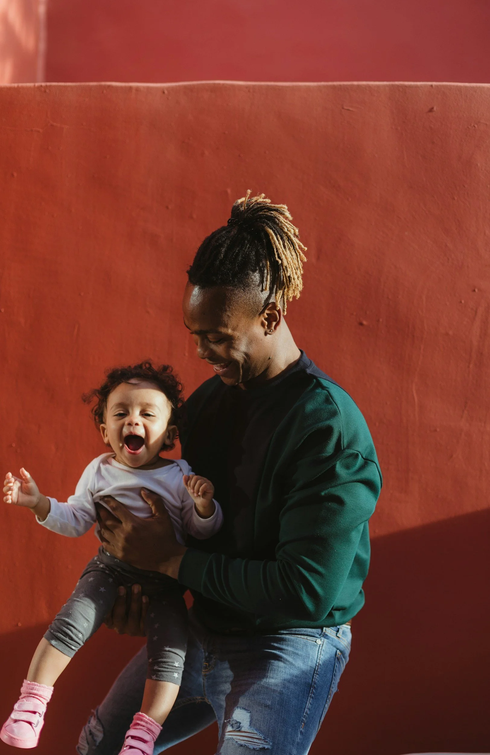 A smiling man with blond dreadlocks holding a young girl with curly hair, both standing in front of a red wall.