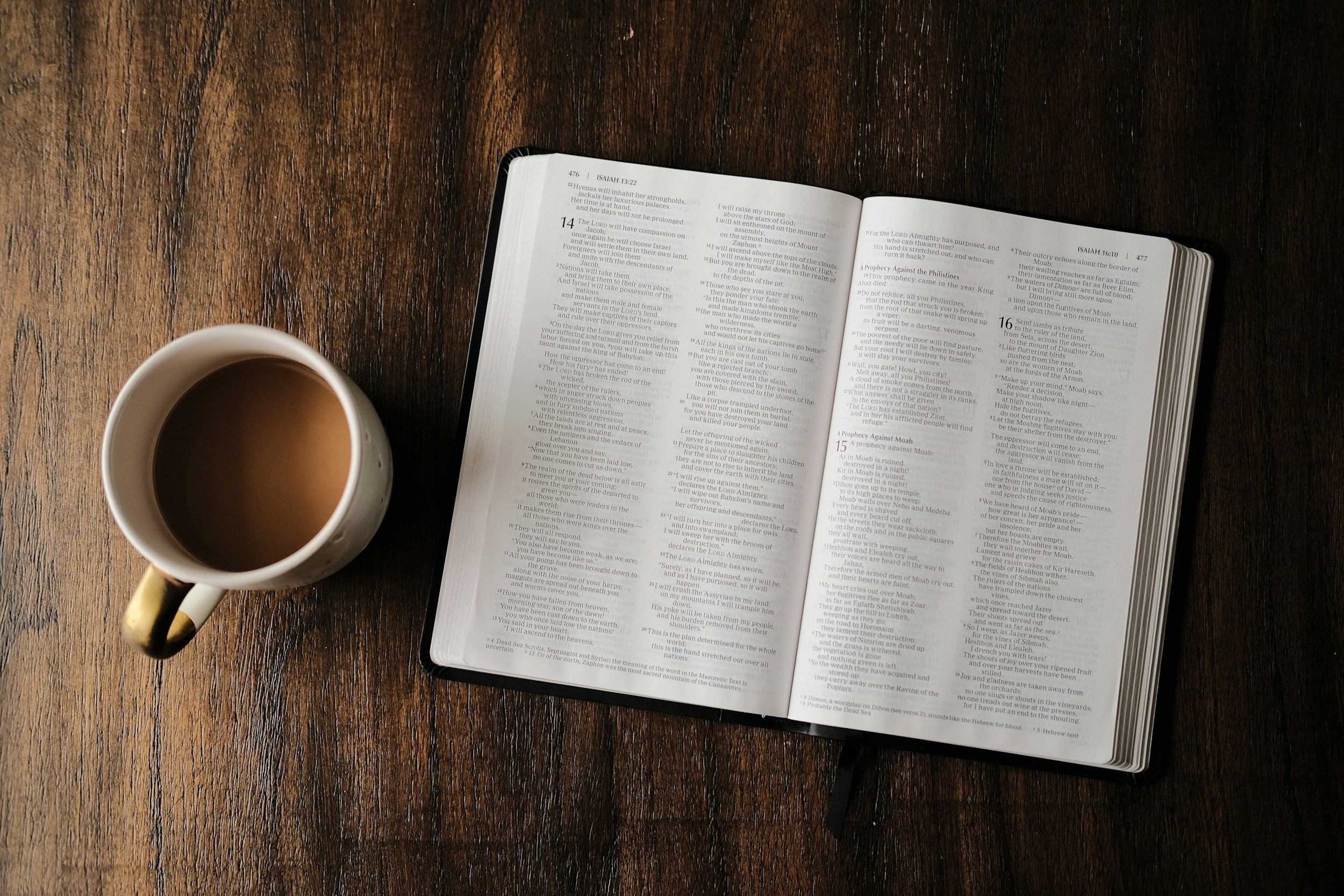 An open Bible on a wooden table next to a white mug filled with coffee.