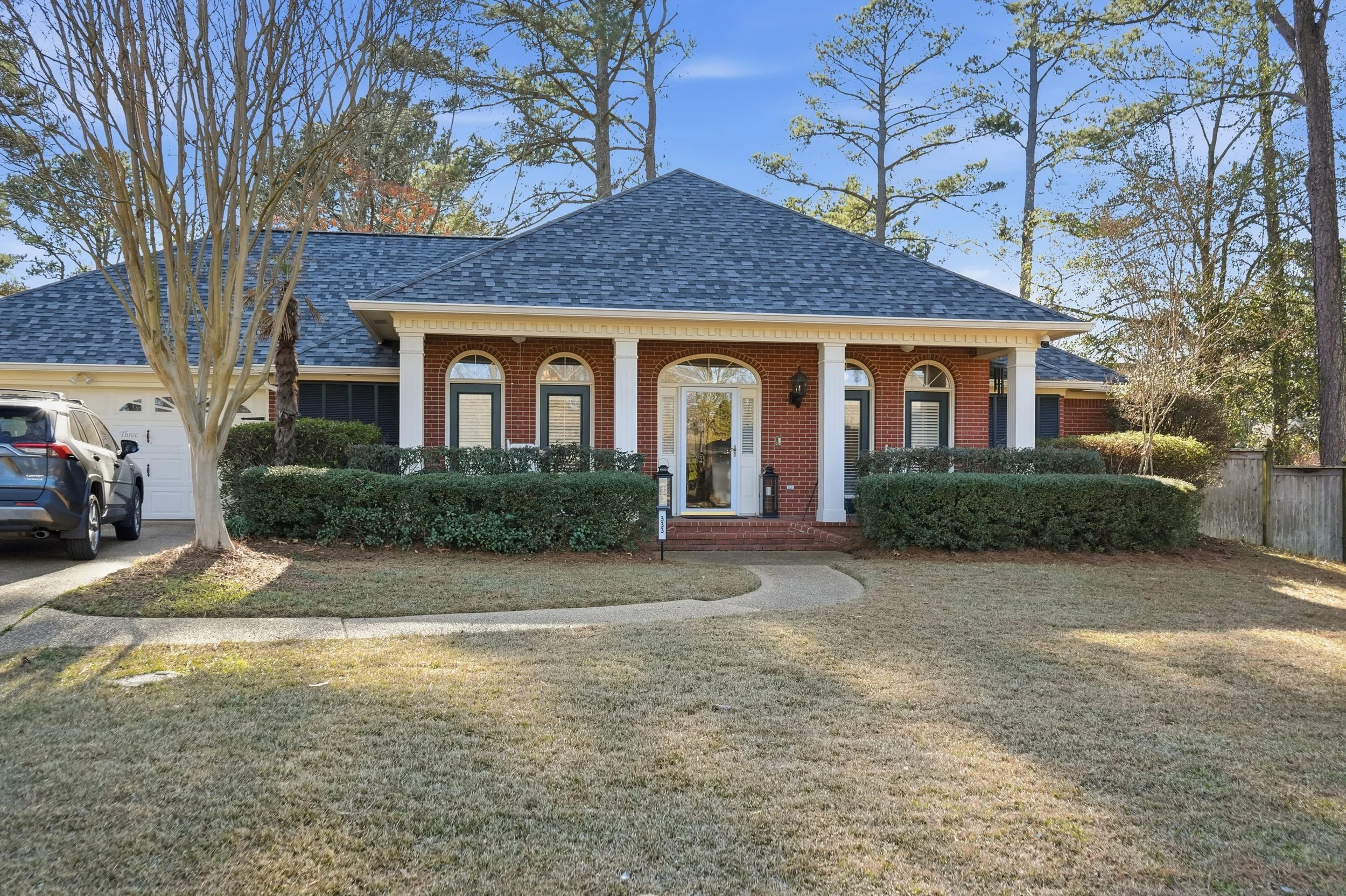 Front view of a brick house with a landscaped yard, concrete pathway, and several windows, surrounded by trees.