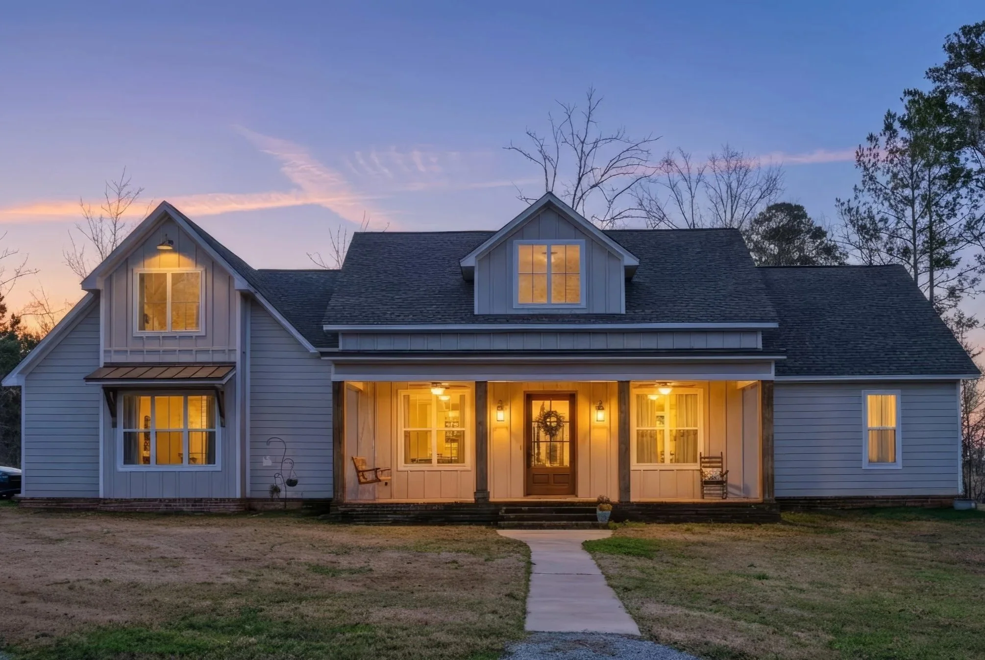 A two-story house with a front porch, lit by interior lights at sunset. The house has white siding, a gabled roof, and several windows, with a concrete pathway leading to the front door.