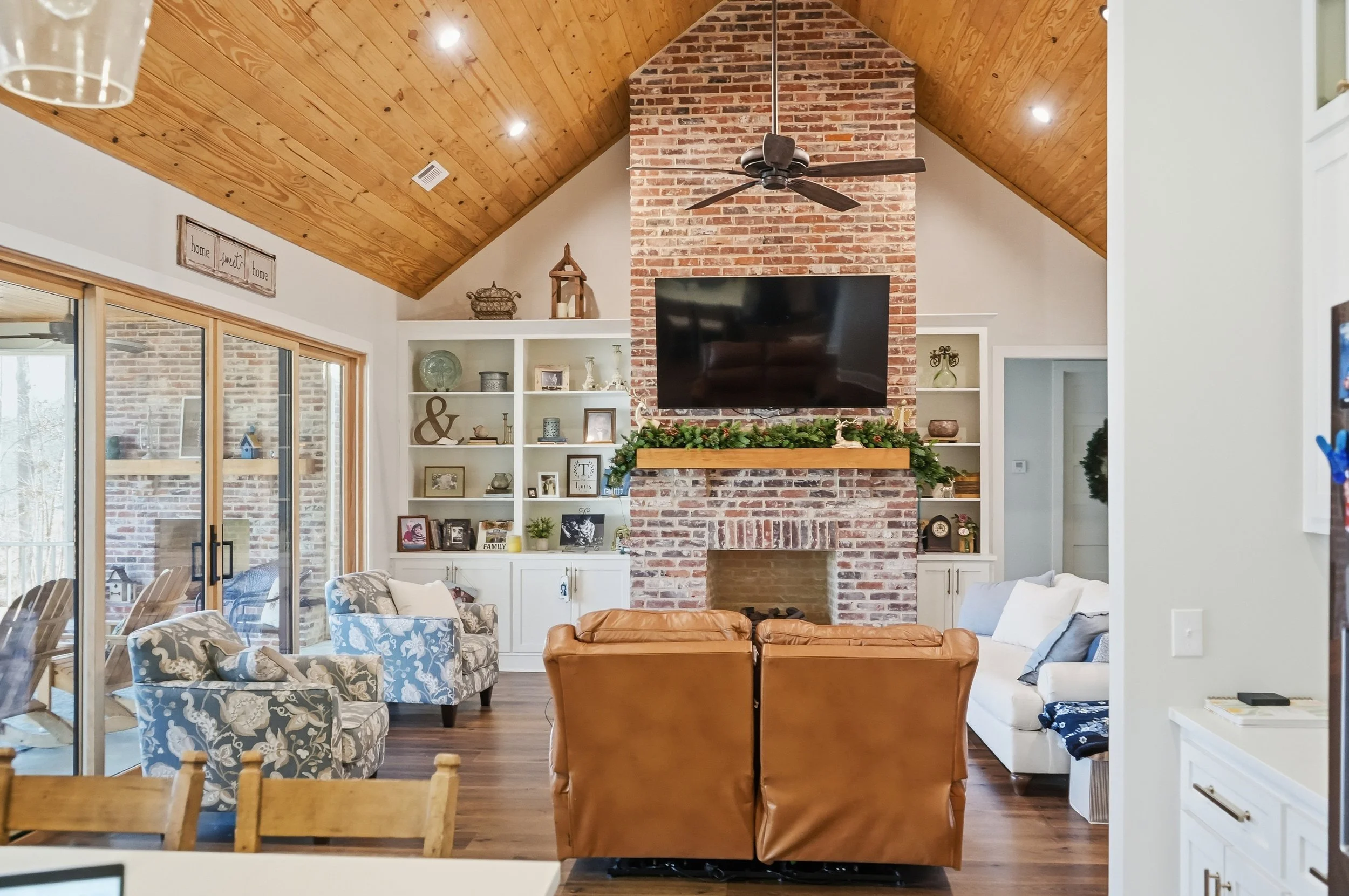 Living room with a brick fireplace, built-in white shelves, a large flat-screen TV, a wooden mantel with greenery, a leather armchair, a patterned armchair, a sofa, and a glass sliding door leading to a patio.