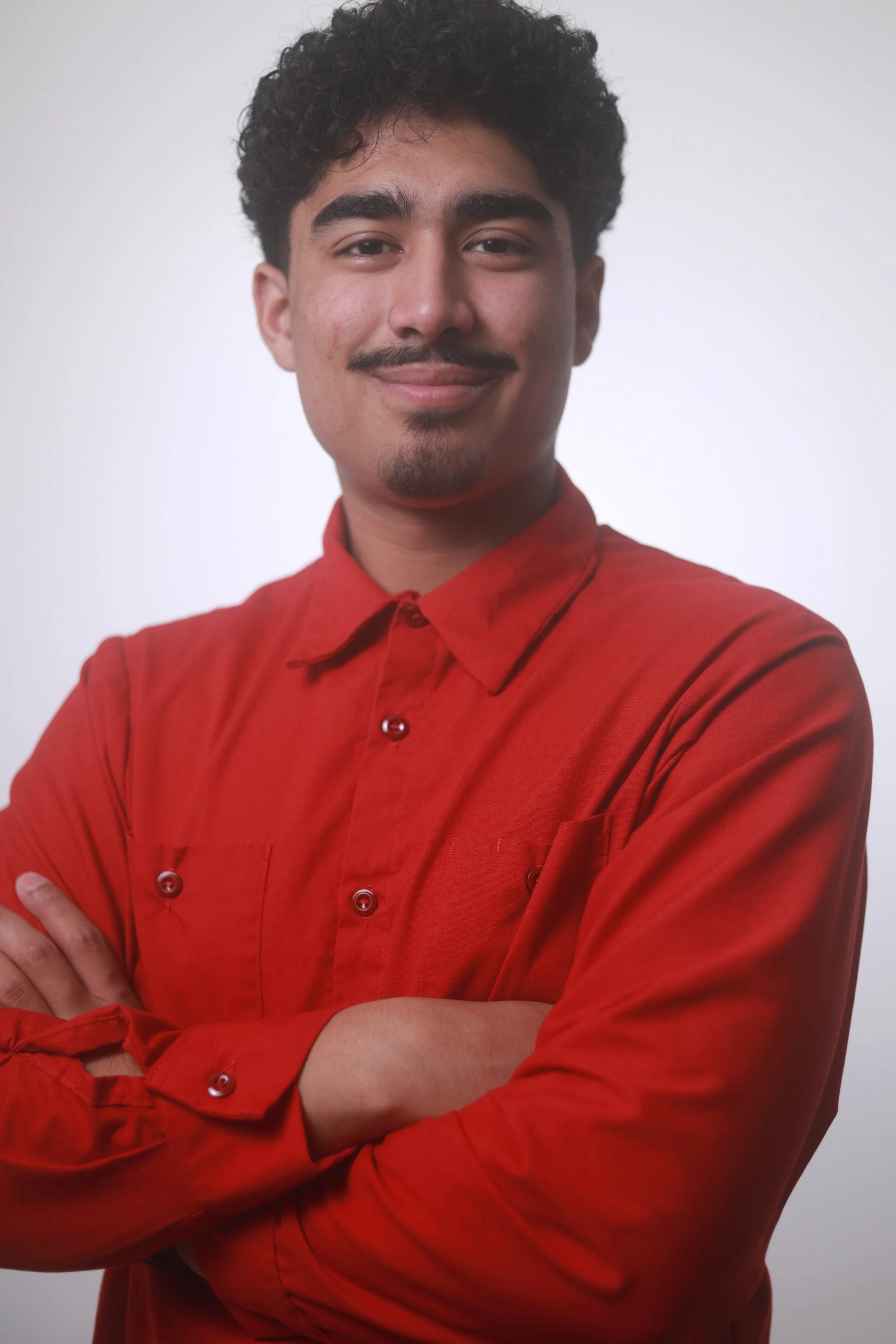 Young man with curly hair wearing a red button-up shirt, smiling and crossing arms against a plain white background.