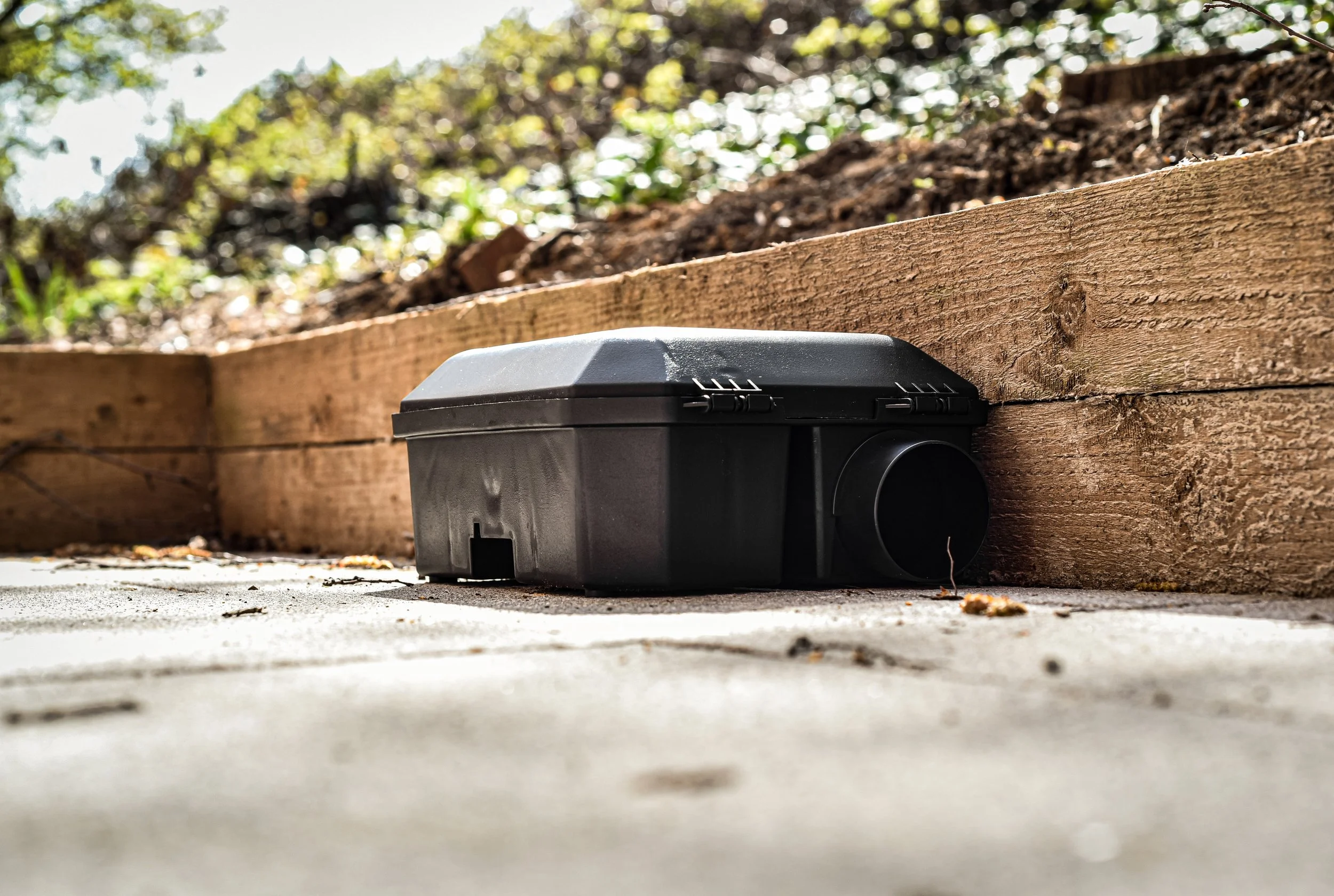 Black rodent bait station on a concrete surface near a wooden retaining wall, with soil and foliage in the background.
