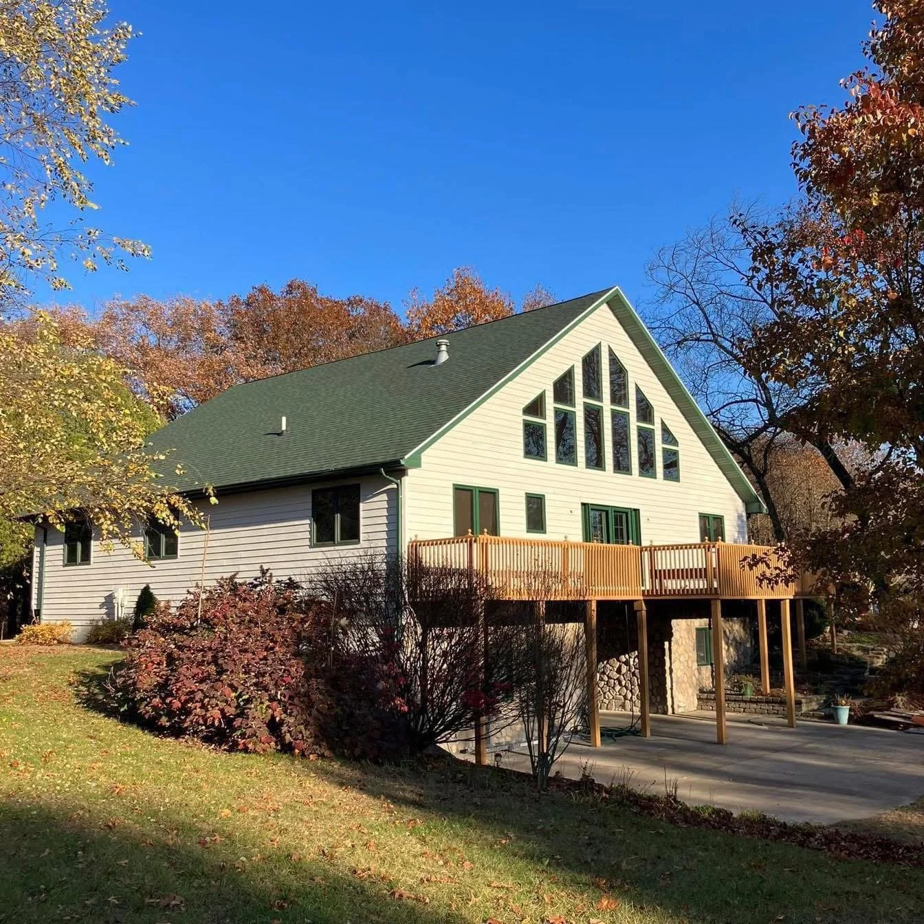 A house with beige siding and green roof, featuring large triangular windows, a balcony, and a paved driveway, surrounded by autumn trees and a grassy lawn under a blue sky.