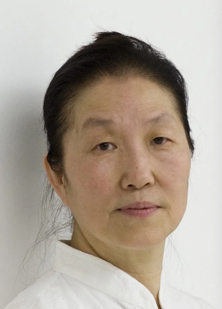 A portrait of an Asian woman with short black hair, wearing a white top, standing against a plain white background.
