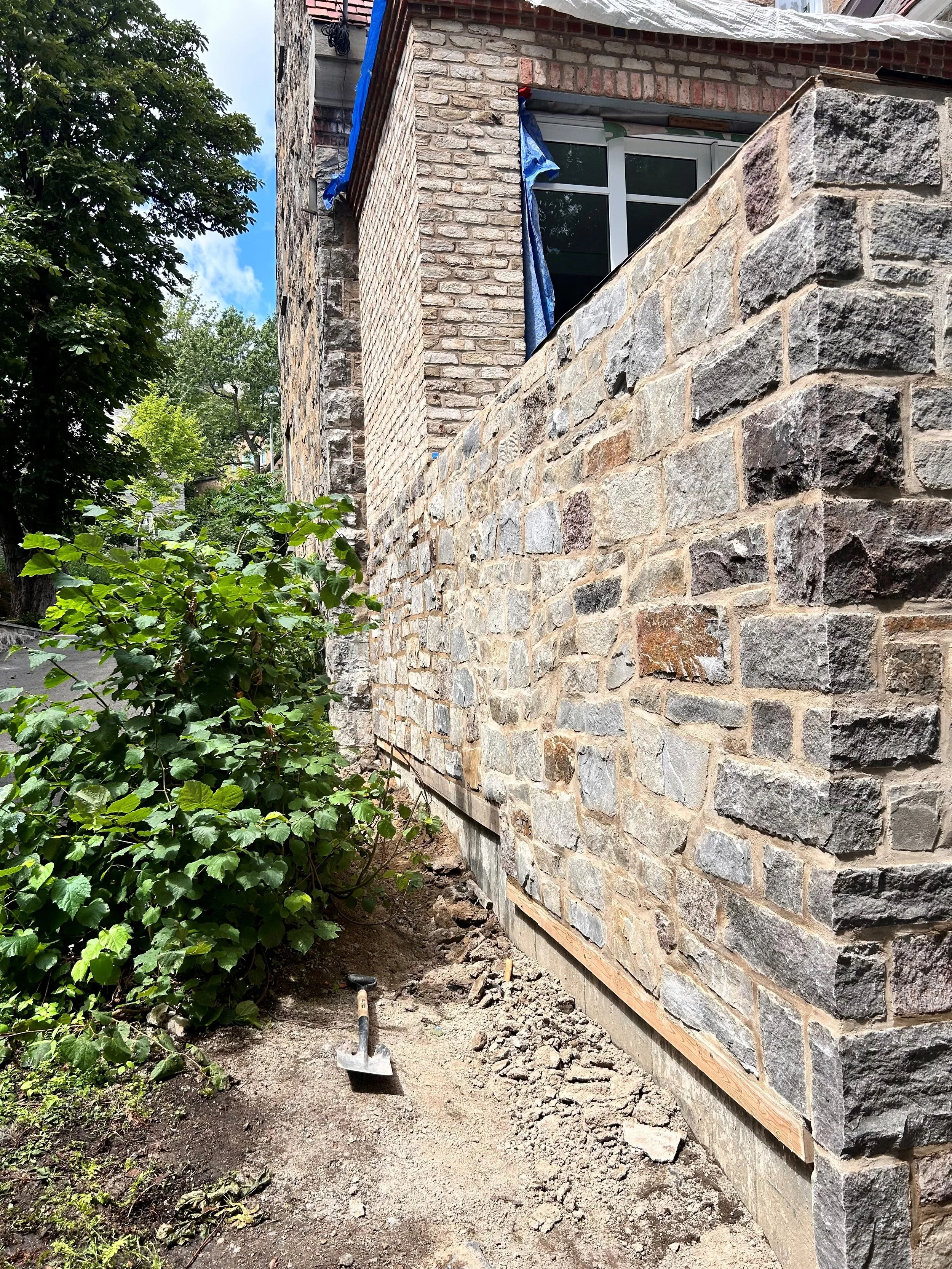 Travaux de construction ou de rénovation avec mur en pierre et outil de chantier posé à terre, côté jardin avec végétation et une maison en arrière-plan, ciel partiellement nuageux.