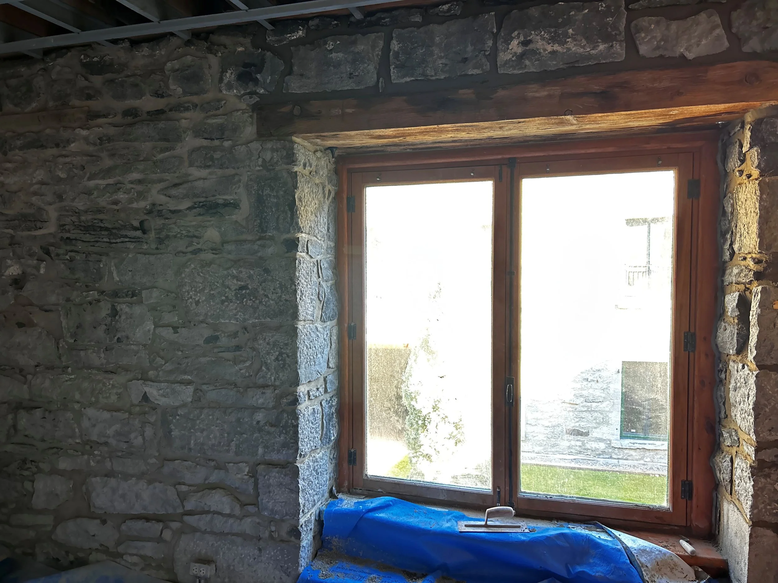 Fenêtre en bois encadrée de pierres dans une pièce en construction ou rénovation, avec un bâche bleue posée en dessous.
