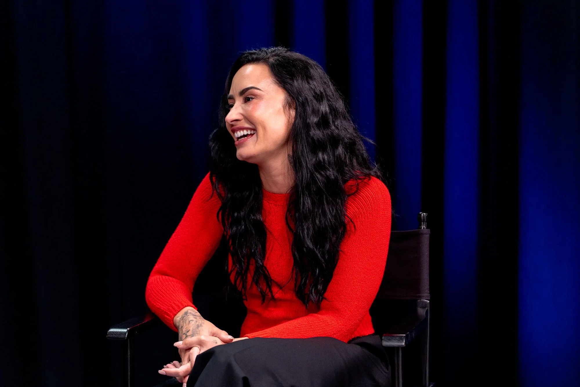Woman with long black hair smiling, wearing a red sweater, sitting on a black chair against a dark blue curtain background.